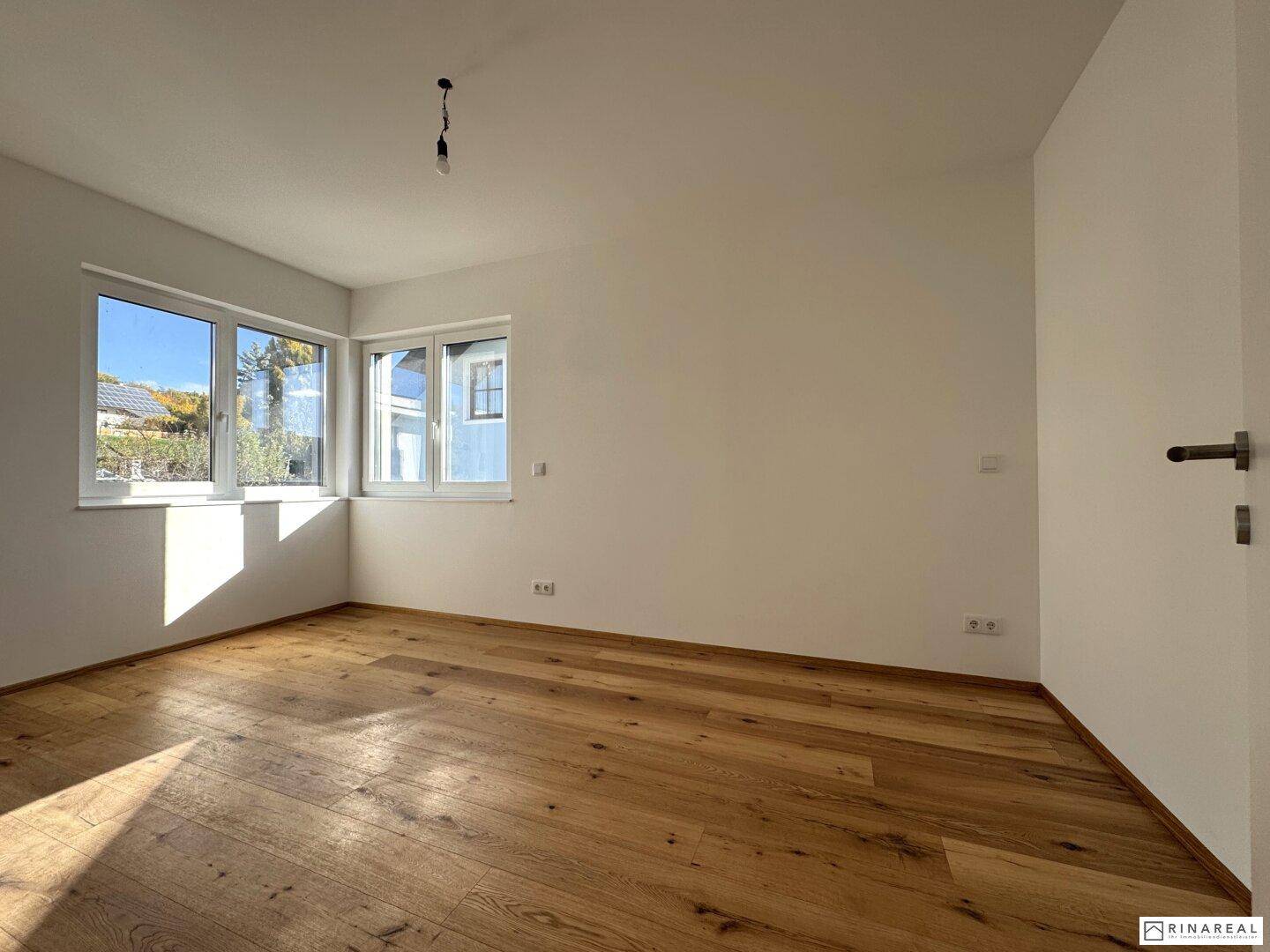 Helles Zimmer mit Holzboden und zwei Fenstern, das viel Sonnenlicht hereinlässt.