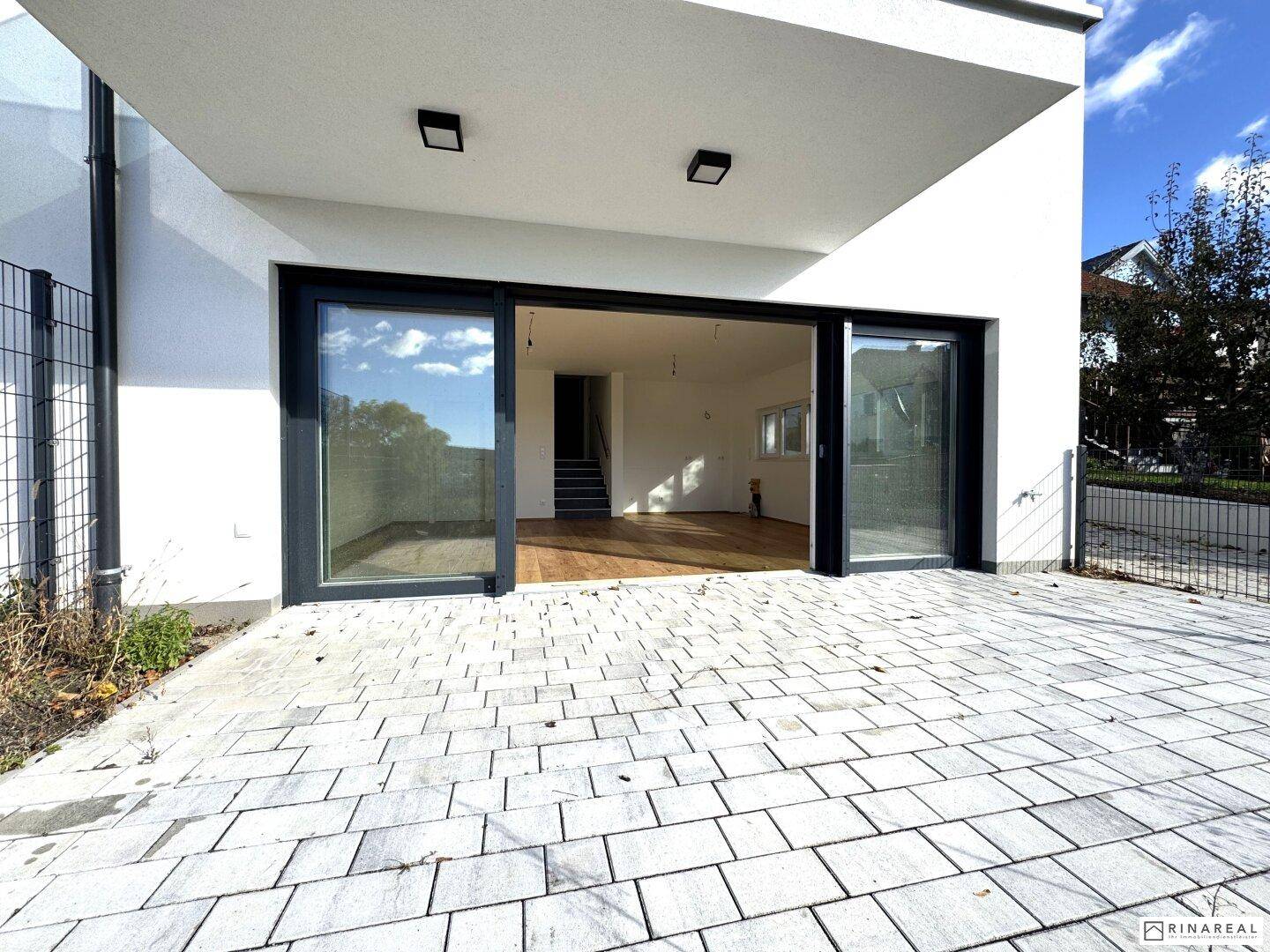 Gepflasterte Terrasse im Erdgeschoss mit Zugang zu einem hellen, leeren Raum durch große Glasschiebetüren.