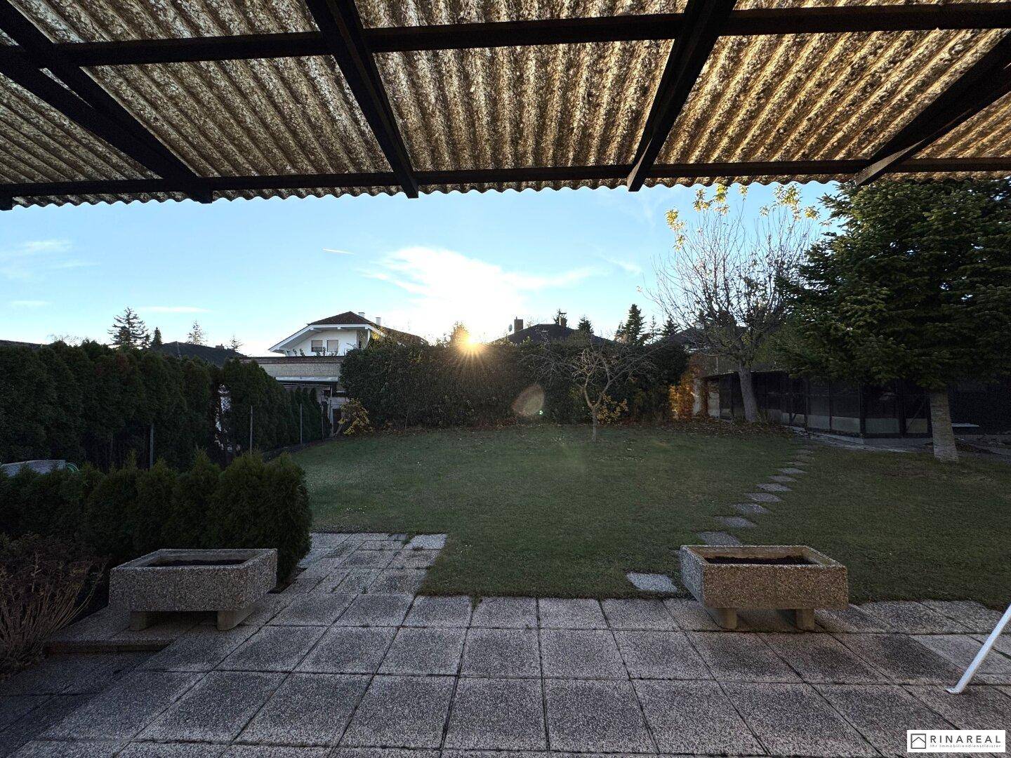 Blick von der überdachten Terrasse in den grünen Garten mit Abendsonne.