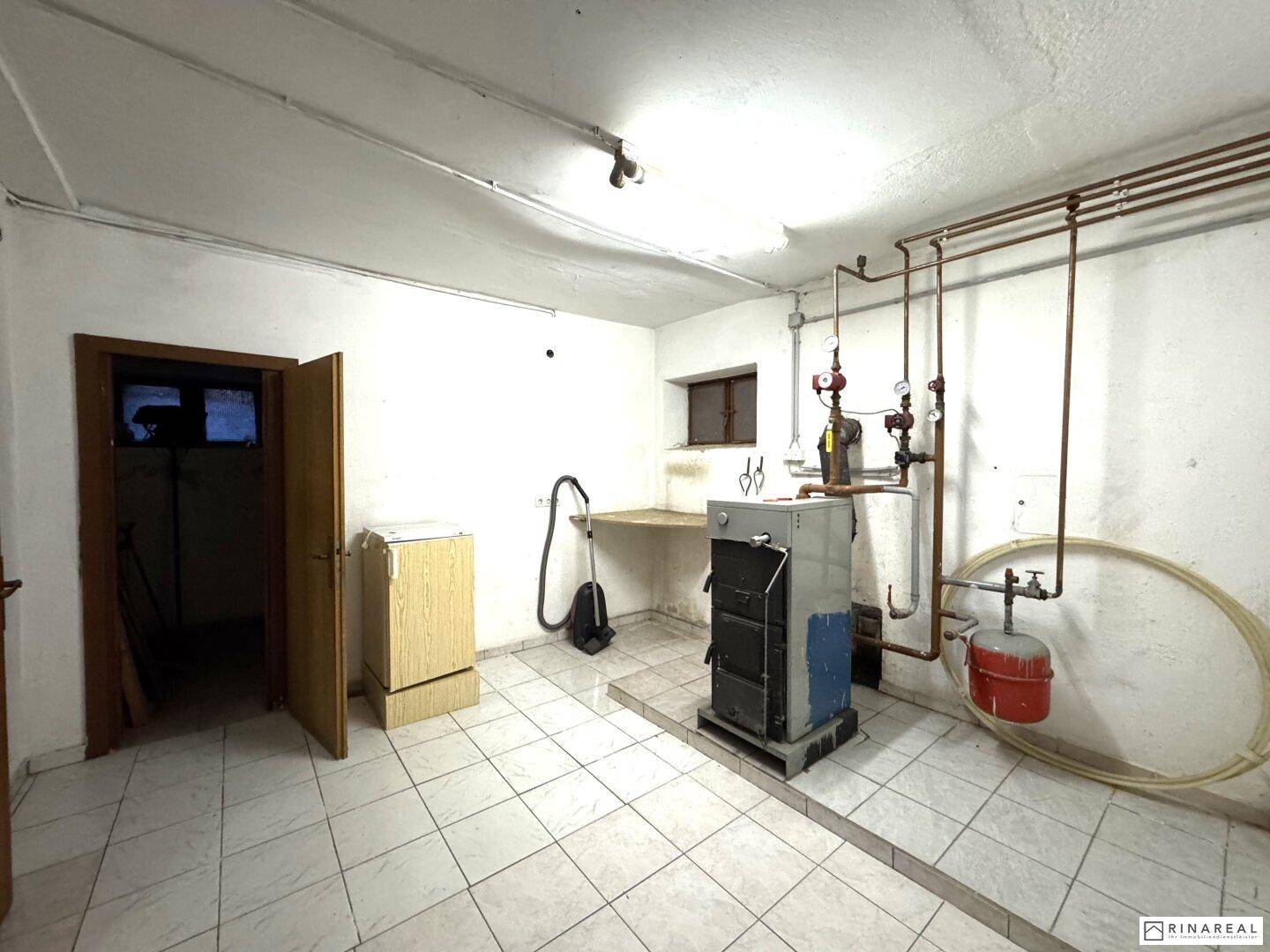Heizungsraum im Keller mit Kessel, Rohren und Fliesenboden.