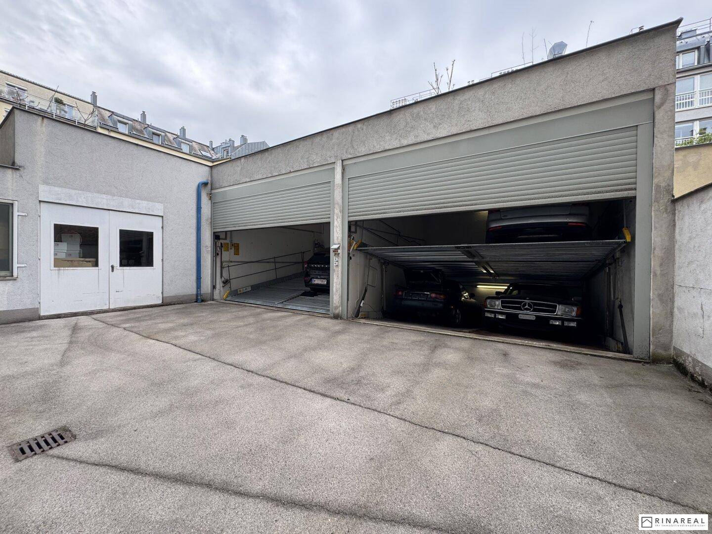 Außenbereich der Garage mit zwei Stellplätzen und angrenzendem Nebengebäude auf einem betonierten Vorplatz.