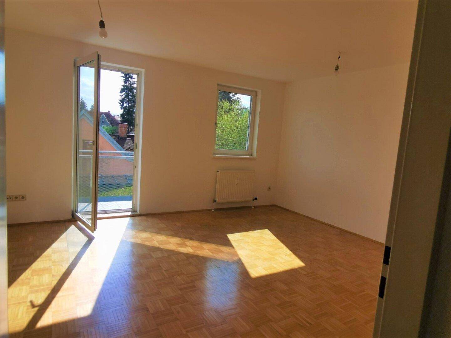 Helles, leeres Zimmer mit Parkettboden, Heizkörper und Zugang zu einem Balkon.