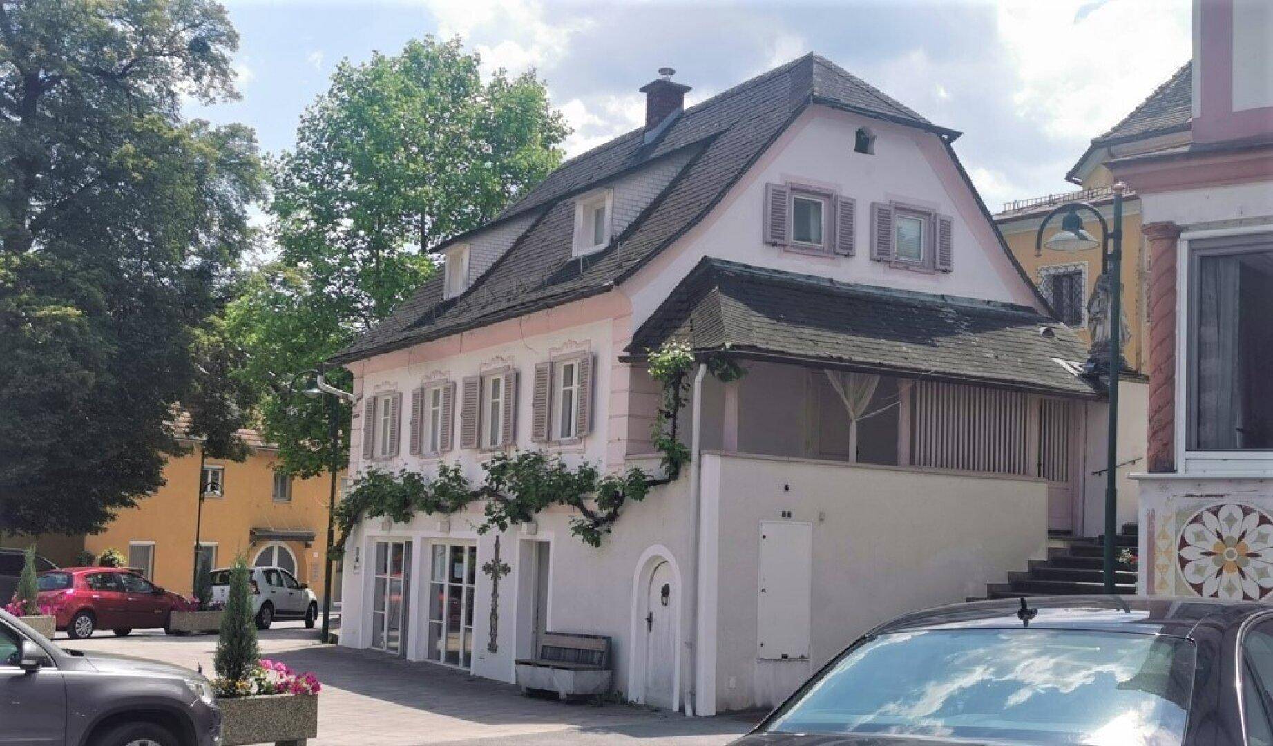 Historisches Gebäude mit traditioneller Fassade, Fensterläden und begrüntem Eingangsbereich an einem sonnigen Tag.
