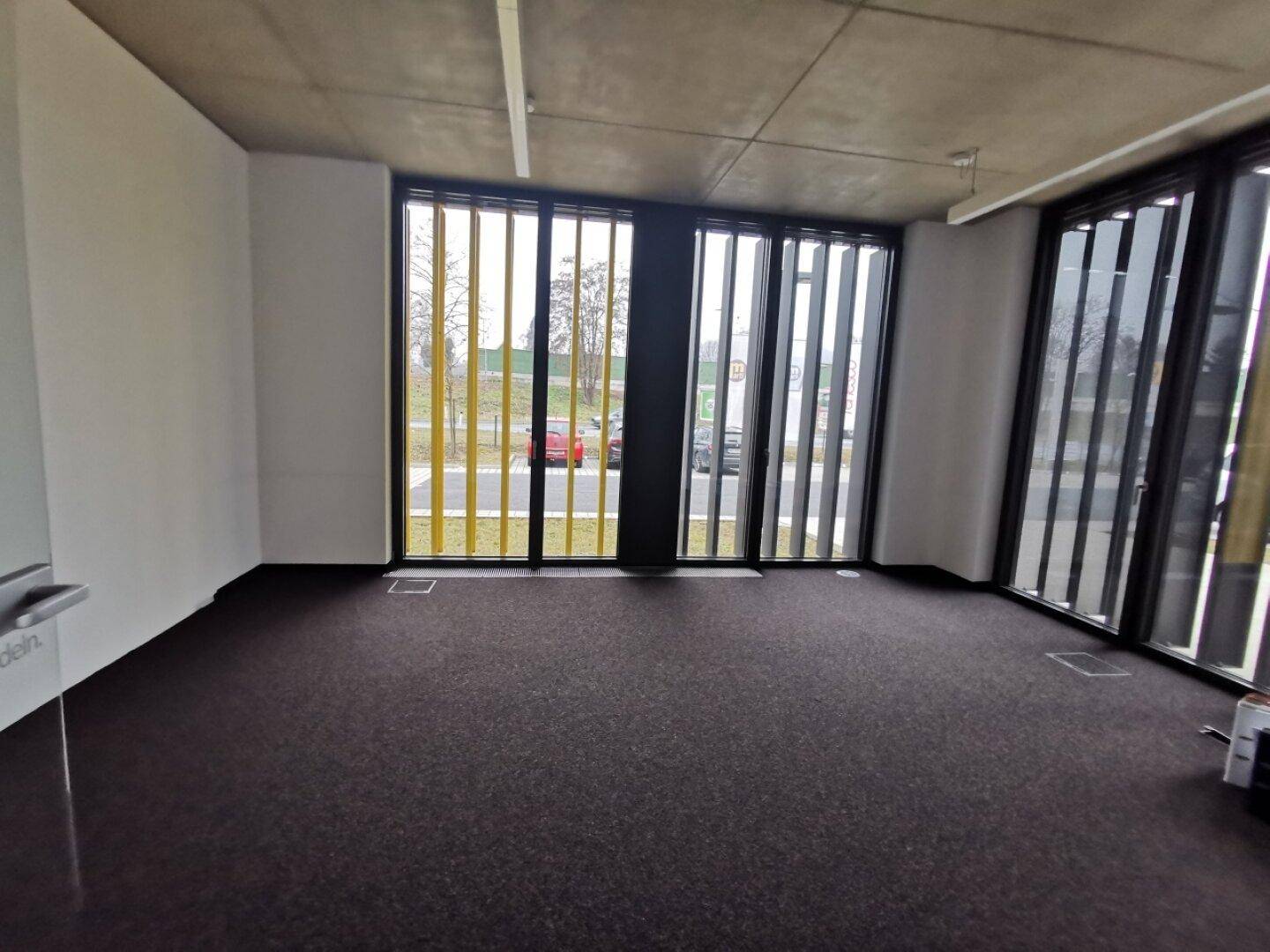 Geräumiger, leerer Büroraum mit dunklem Teppichboden und großen Fenstern mit gelben Lamellen.