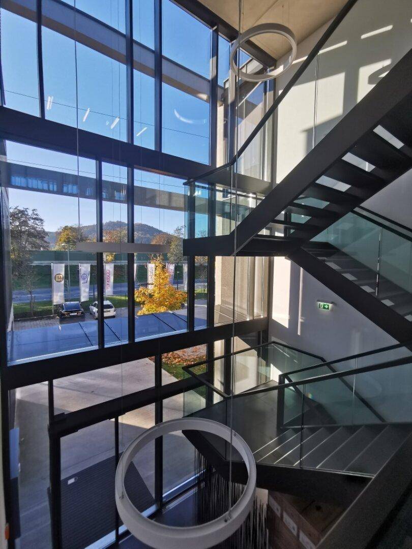 Beeindruckendes Atrium mit moderner Treppe, Glasgeländern und großen Fenstern mit Blick ins Grüne.