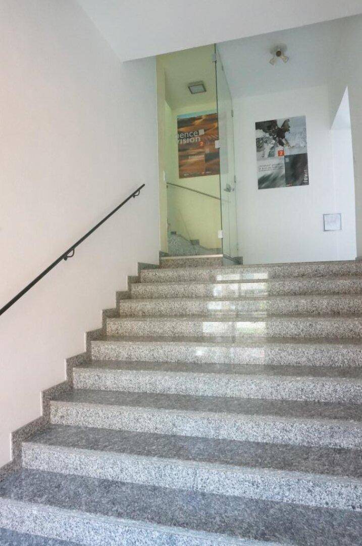 Heller Treppenaufgang mit Granitstufen und einer modernen Glasabtrennung, führt zu den oberen Etagen.