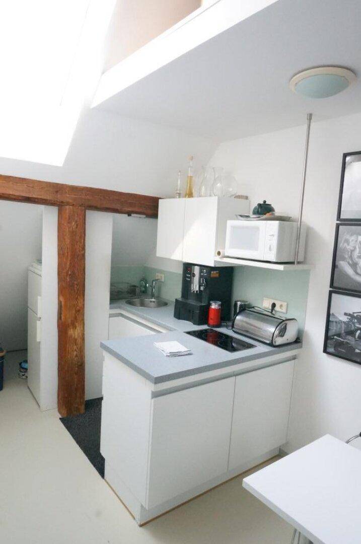 Kleine, moderne Küchenzeile mit weißen Schränken, Mikrowelle und Kaffeemaschine unter einem Dachfenster.