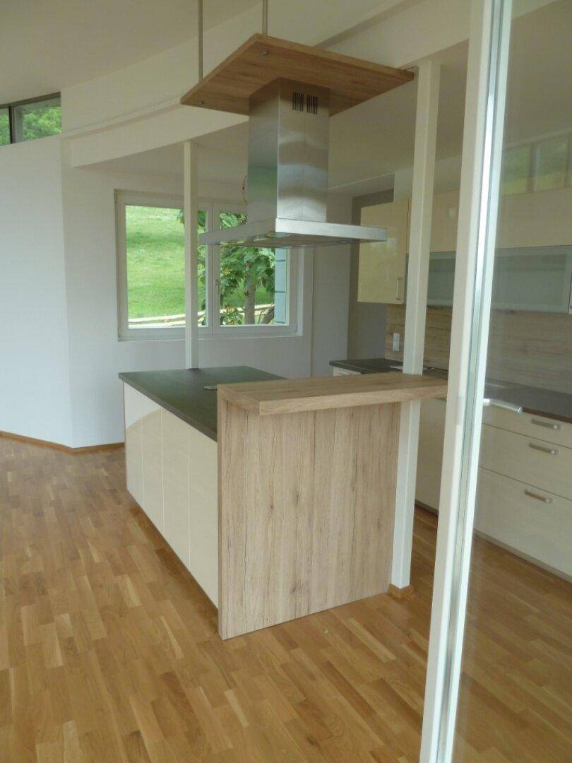 Moderne Küche mit Kochinsel, Dunstabzugshaube und Holzboden, offen zum Wohnbereich.