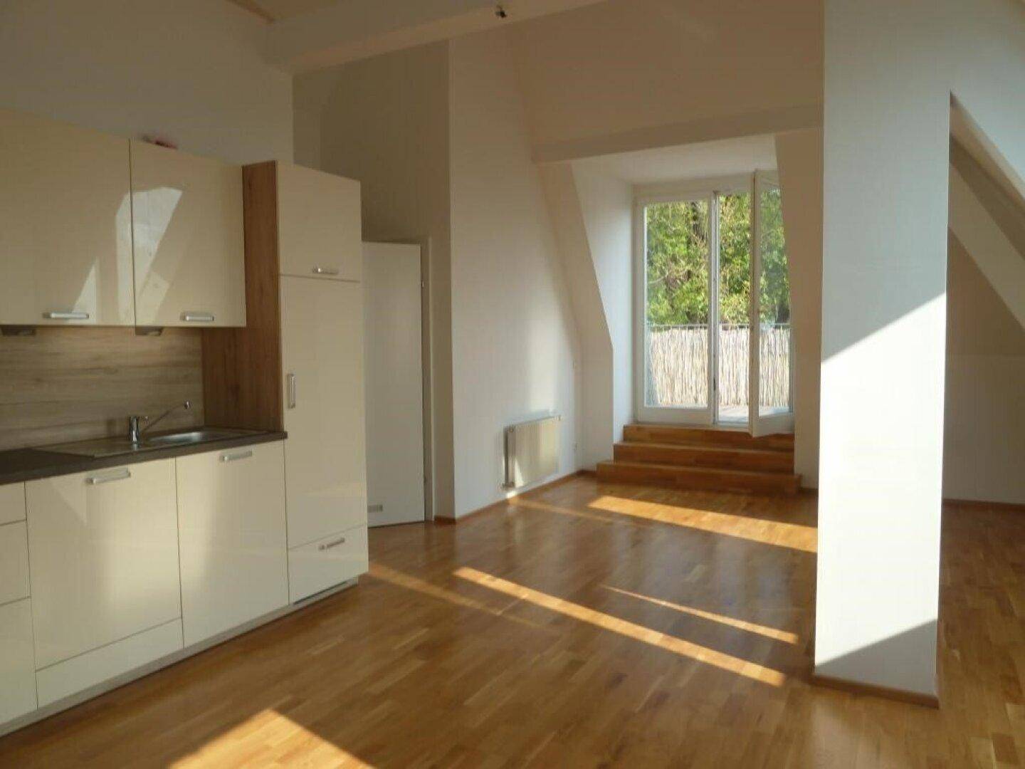 Heller Wohnbereich im Dachgeschoss mit Holzboden, kleiner Küchenzeile und Zugang zu einem Balkon.
