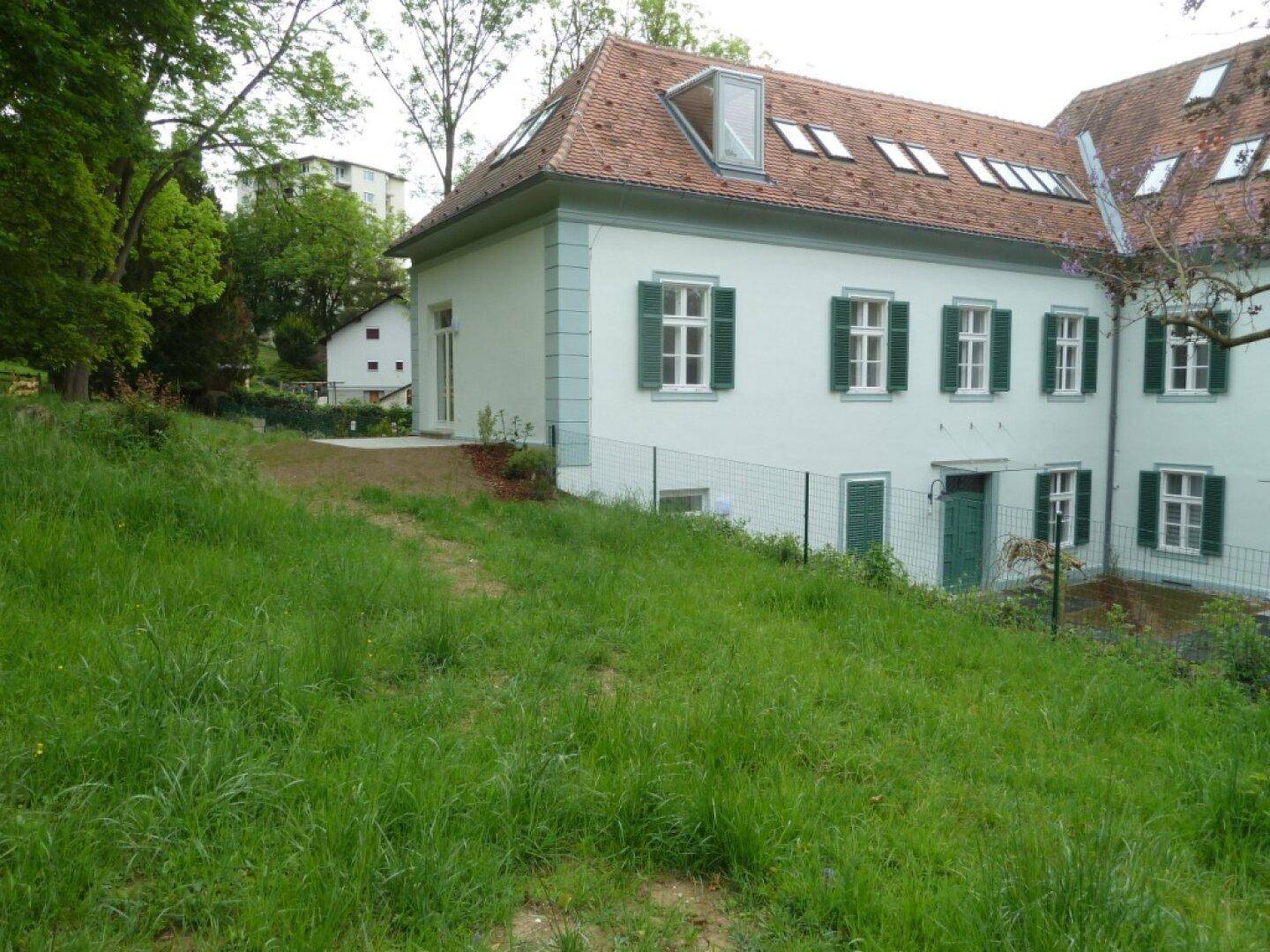 Hellgrünes Gebäude mit Satteldach und grünen Fensterläden, umgeben von einer großen Rasenfläche.