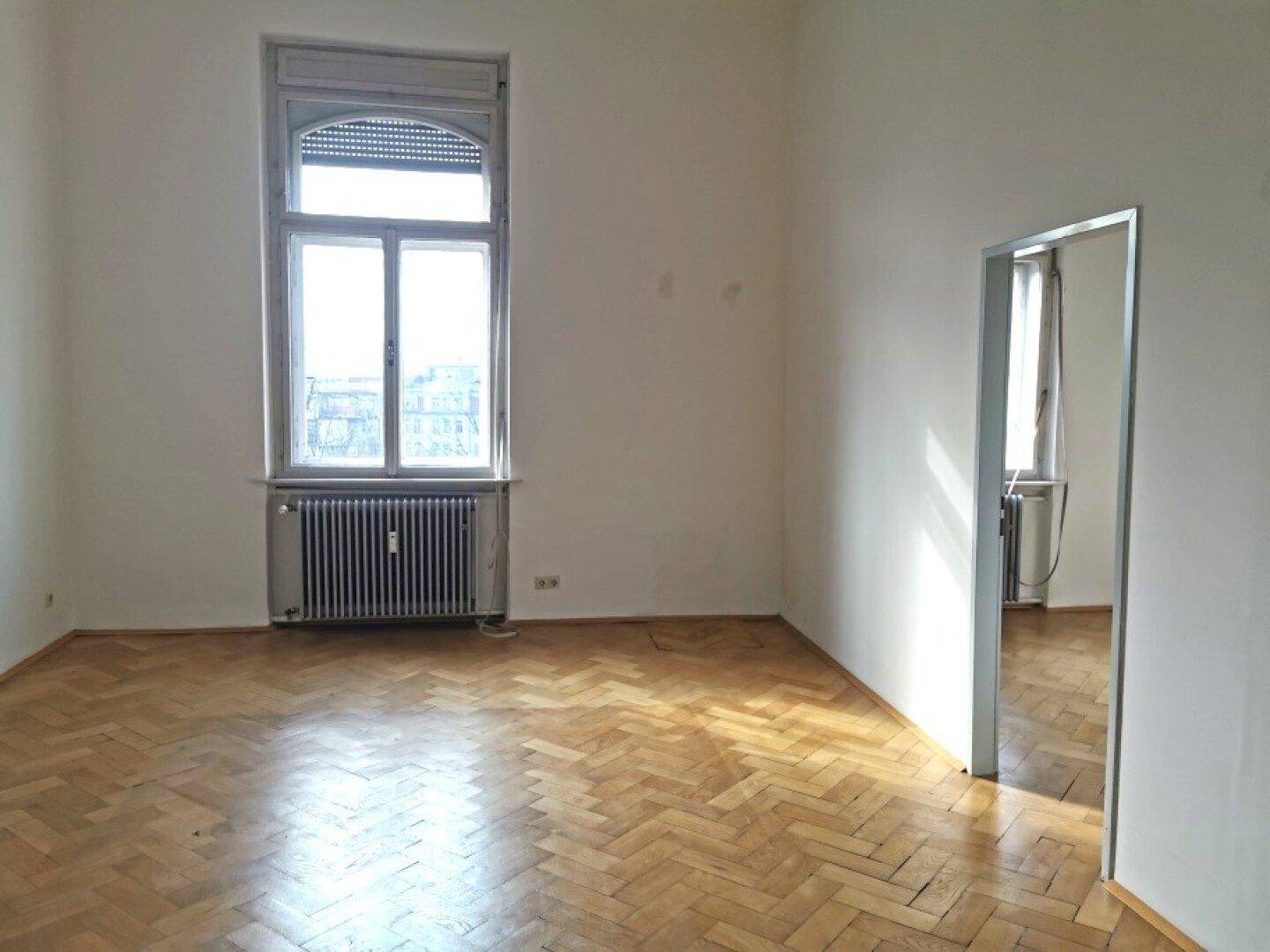 Heller Raum mit Fischgrätparkett, großem Fenster mit Rollladen und einem Spiegel an der Wand.