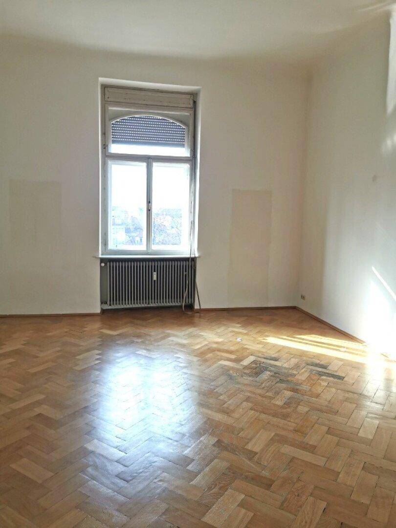 Großer, heller Raum mit Fischgrätparkett und einem Fenster mit Rollladen, das Tageslicht spendet.