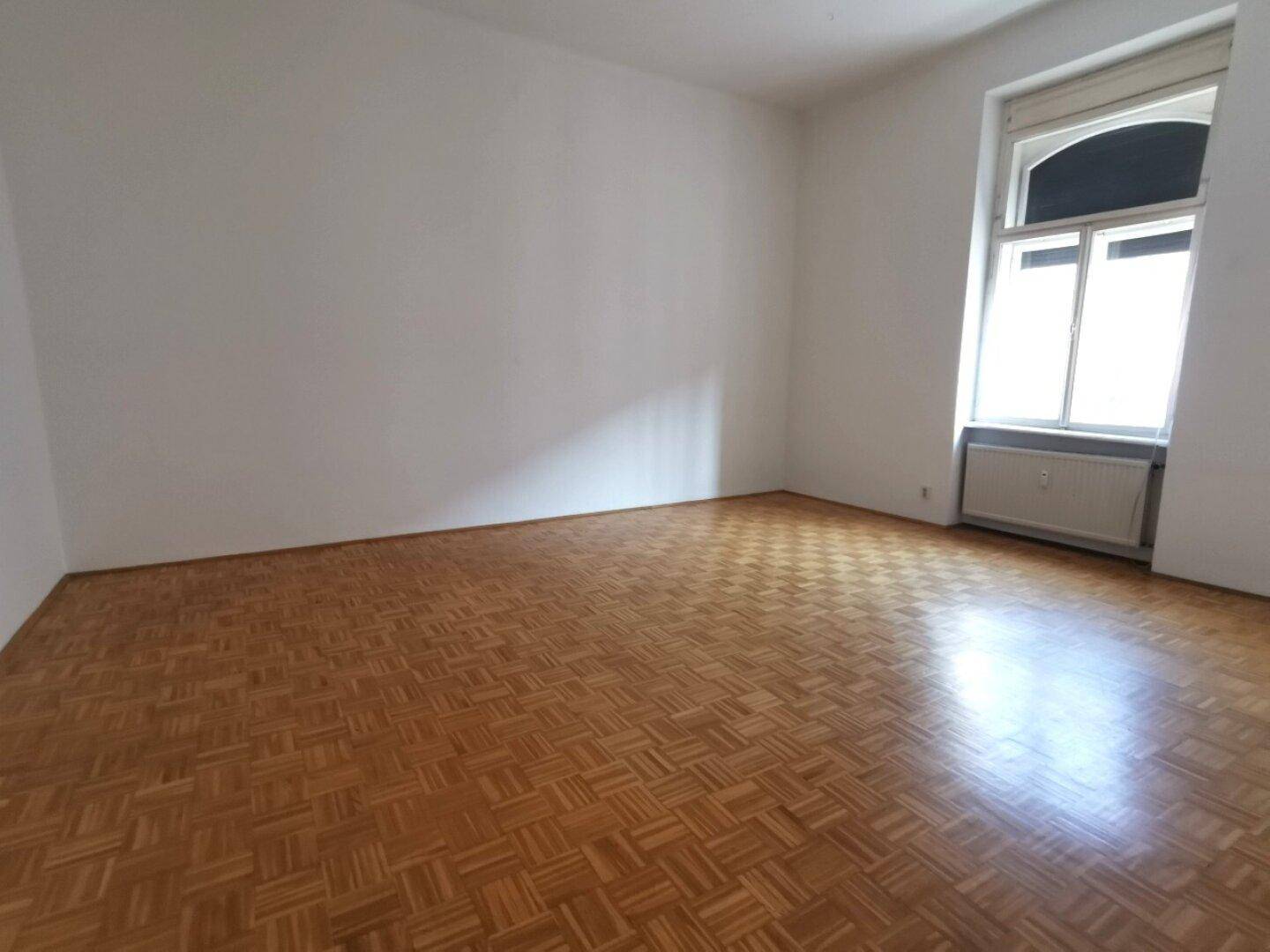 Geräumiger, leerer Raum mit Parkettboden und einem großen Fenster, das viel Tageslicht hereinlässt.