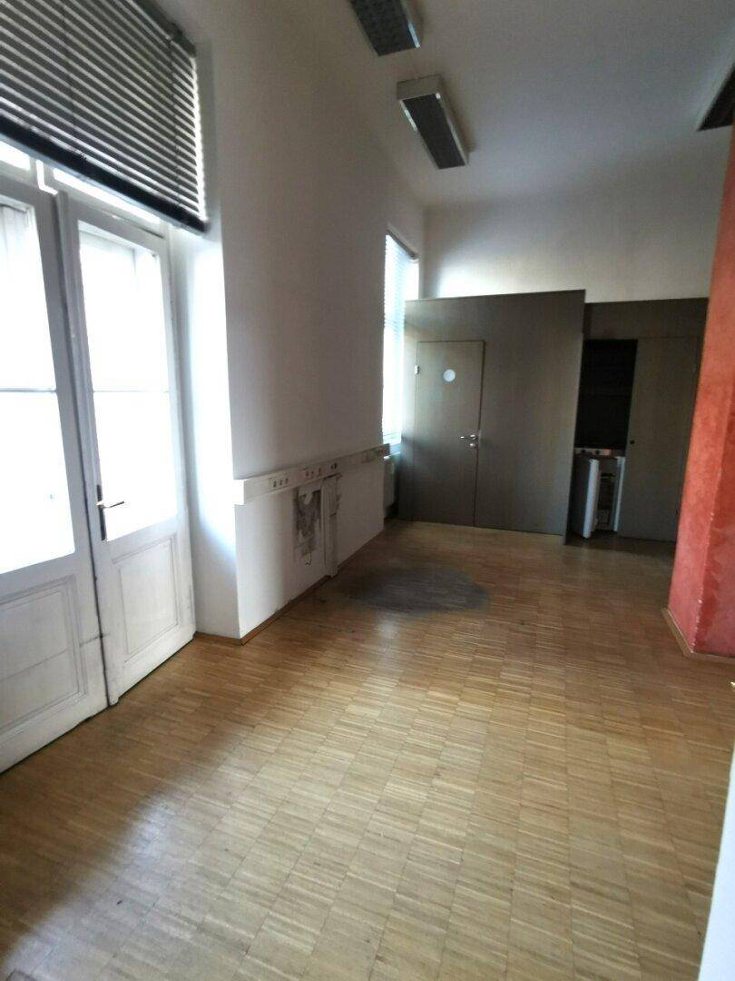 Großer Raum mit Holzboden, Doppeltüren und Leuchtstoffröhren an der Decke, ideal für ein Büro.