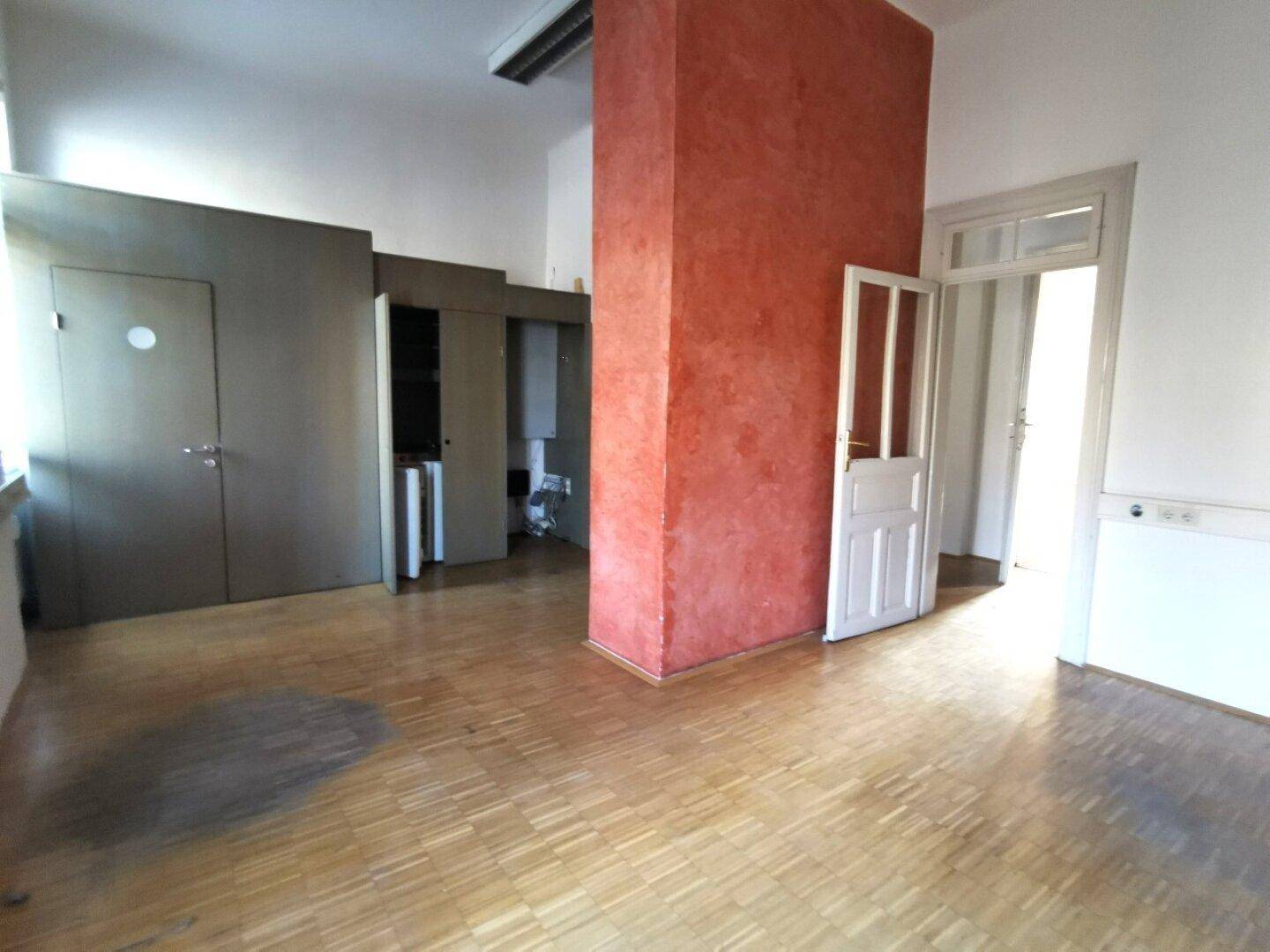 Geräumiger Raum mit Holzboden, roter Säule und mehreren Türen, geeignet als Bürofläche.