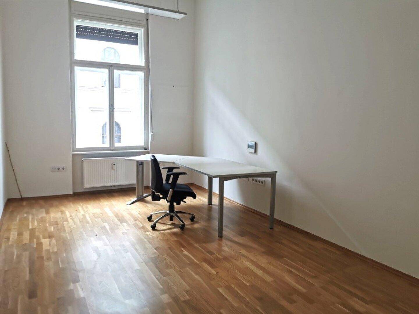Büro mit Holzboden, großem Fenster, Schreibtisch und Bürostuhl, gut beleuchtet.