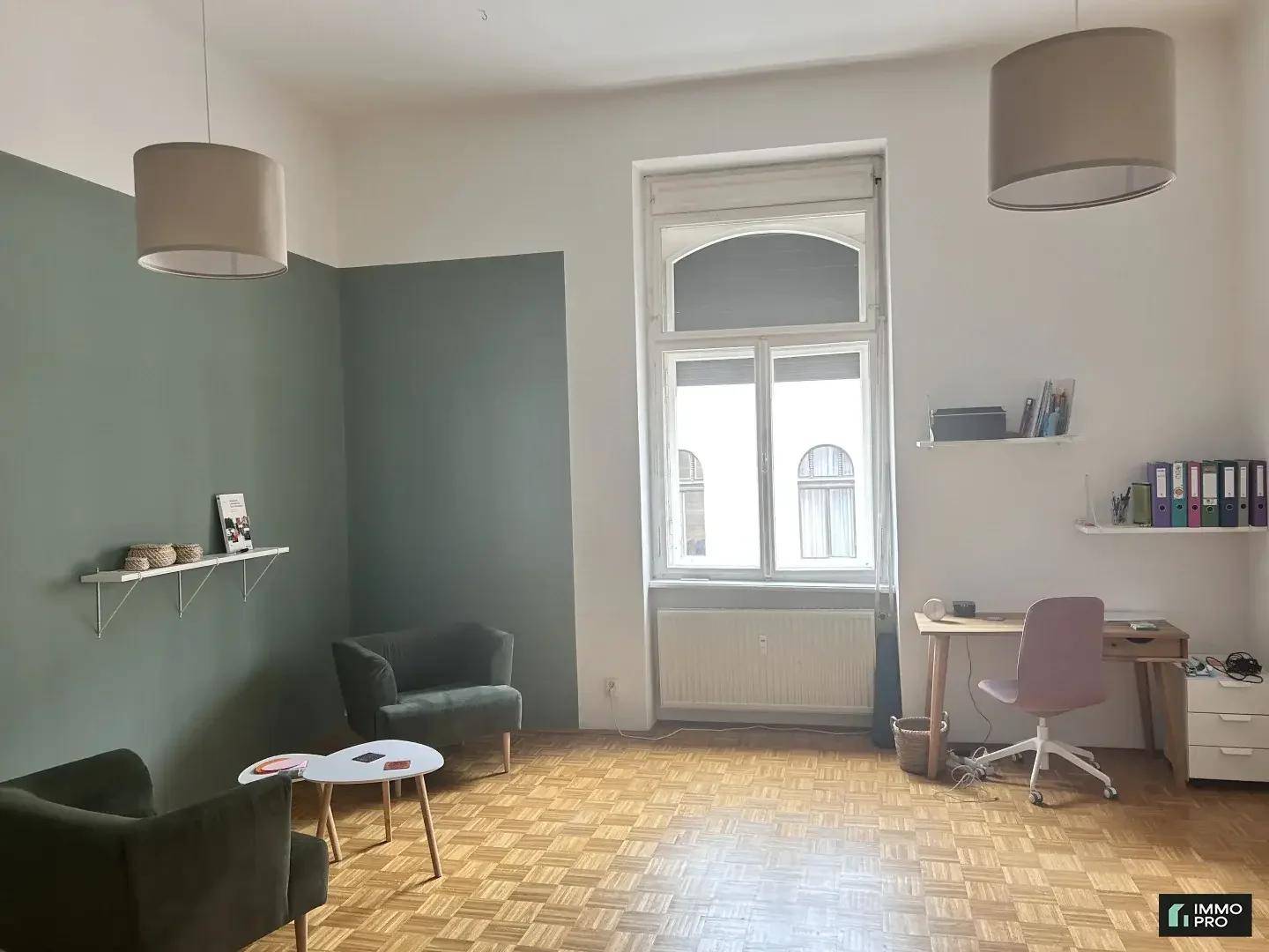 Heller Wohnraum mit Parkettboden, grünen Wänden und einem Arbeitsbereich am großen Fenster.