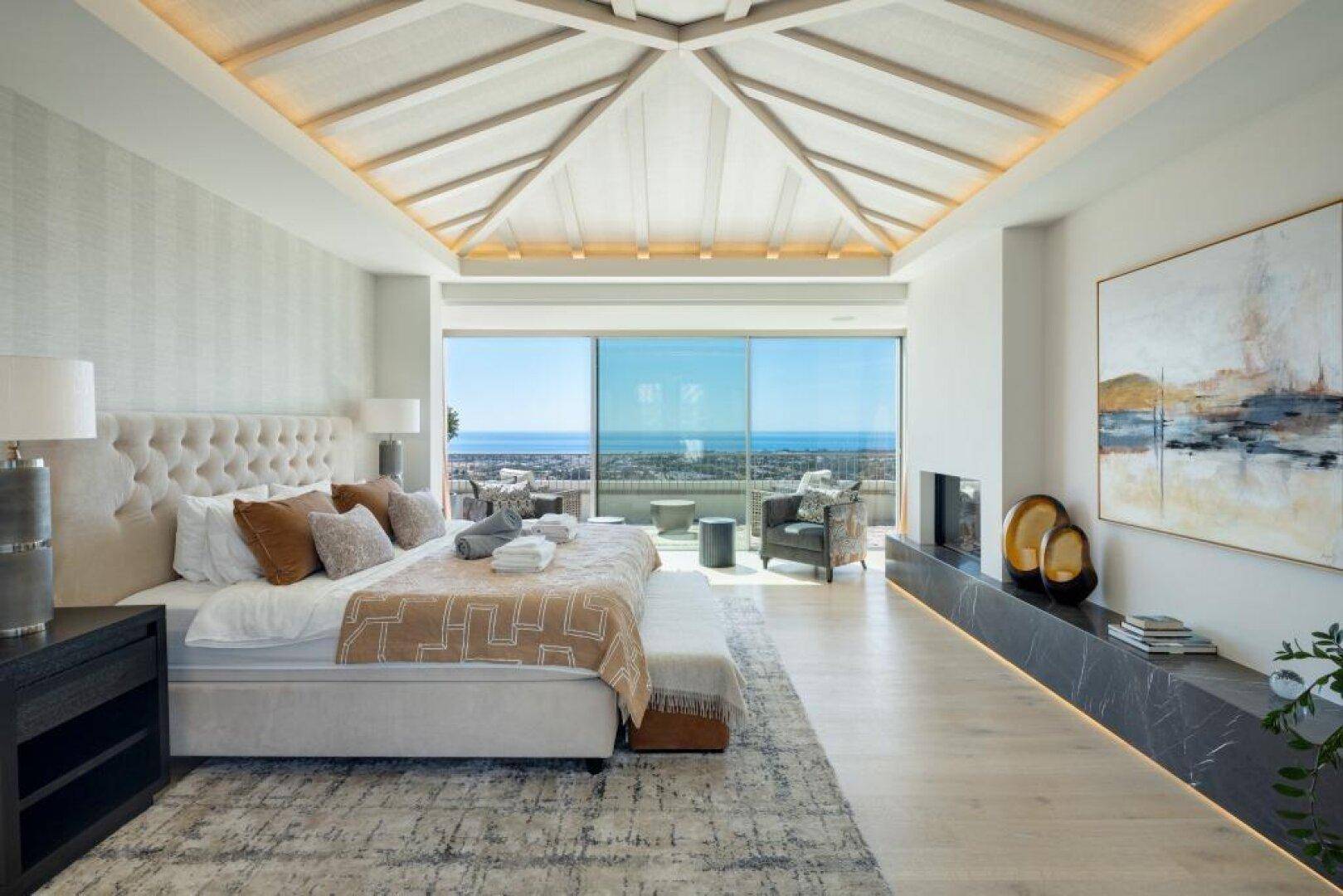 Geräumiges Schlafzimmer mit hoher Decke, Kamin und Meerblick durch große Fenster.
