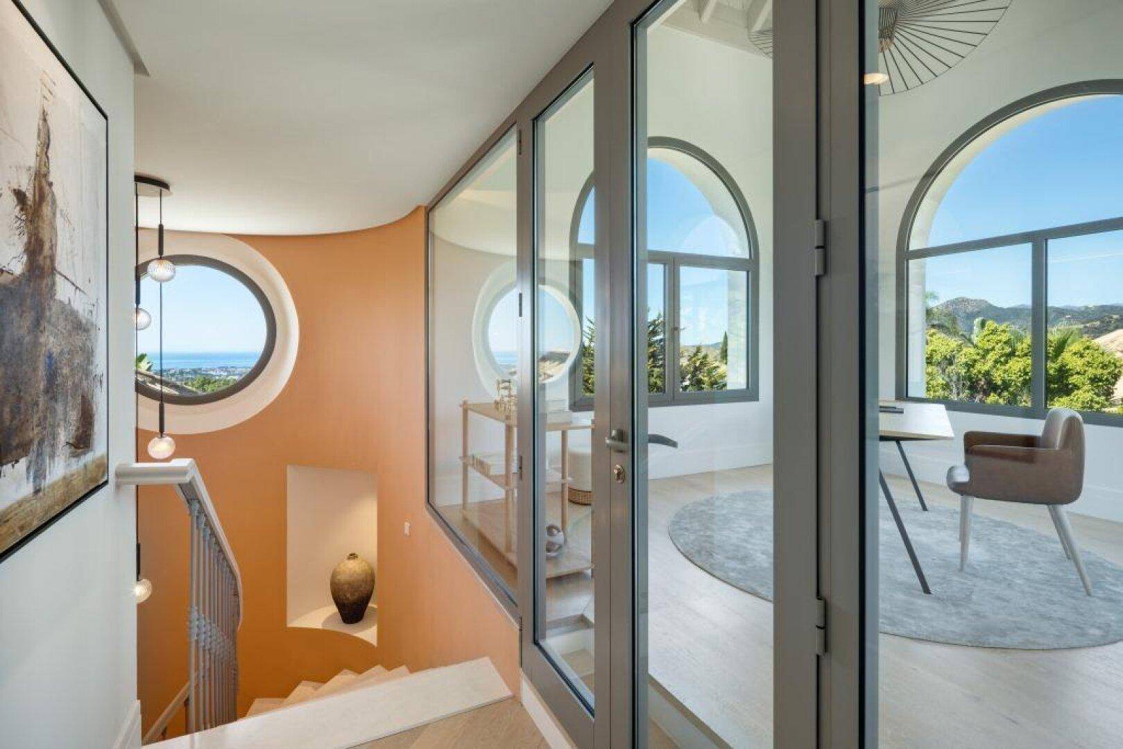 Heller Treppenaufgang mit runden Fenstern und Blick auf die Landschaft und das Meer.