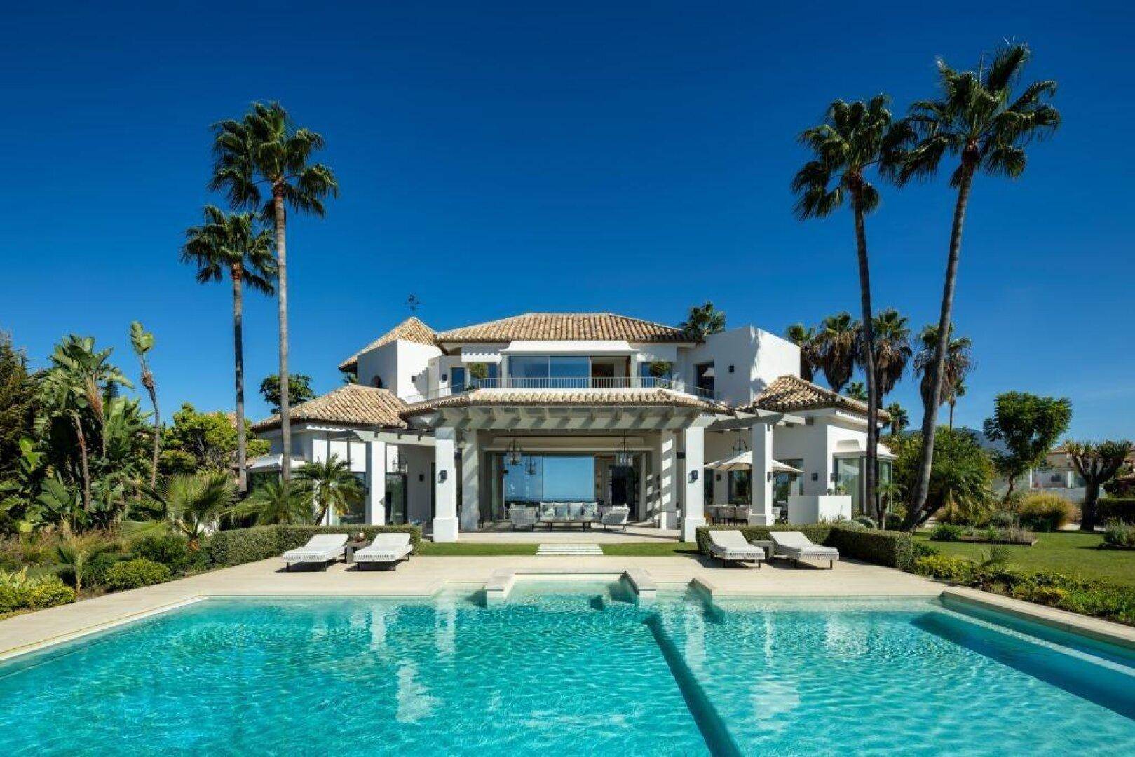 Prächtige Villa mit großem Poolbereich, Palmen und weitläufigem Garten unter blauem Himmel.
