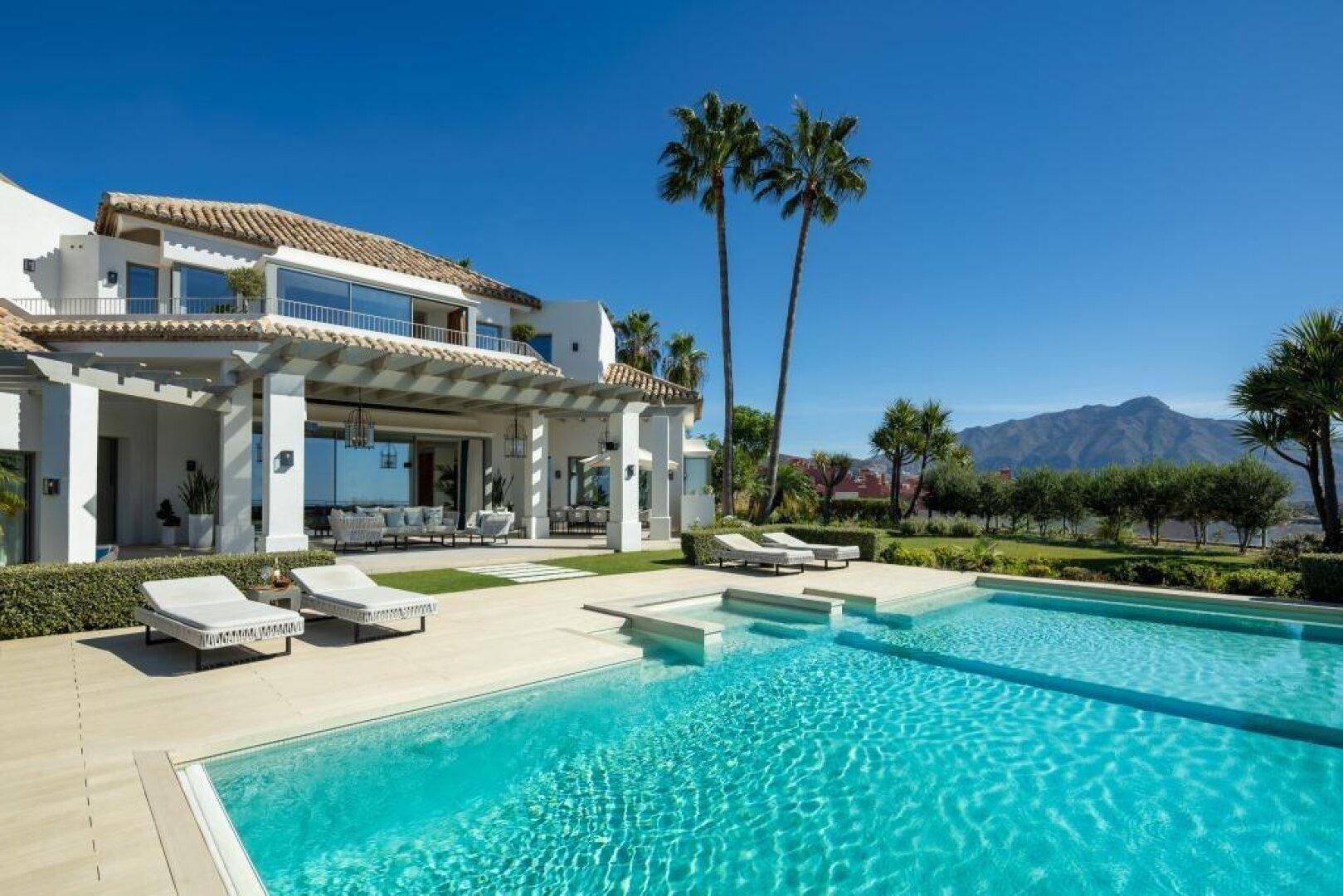 Luxuriöse Villa mit großem Pool, Palmen und beeindruckendem Bergblick unter strahlendem Himmel.