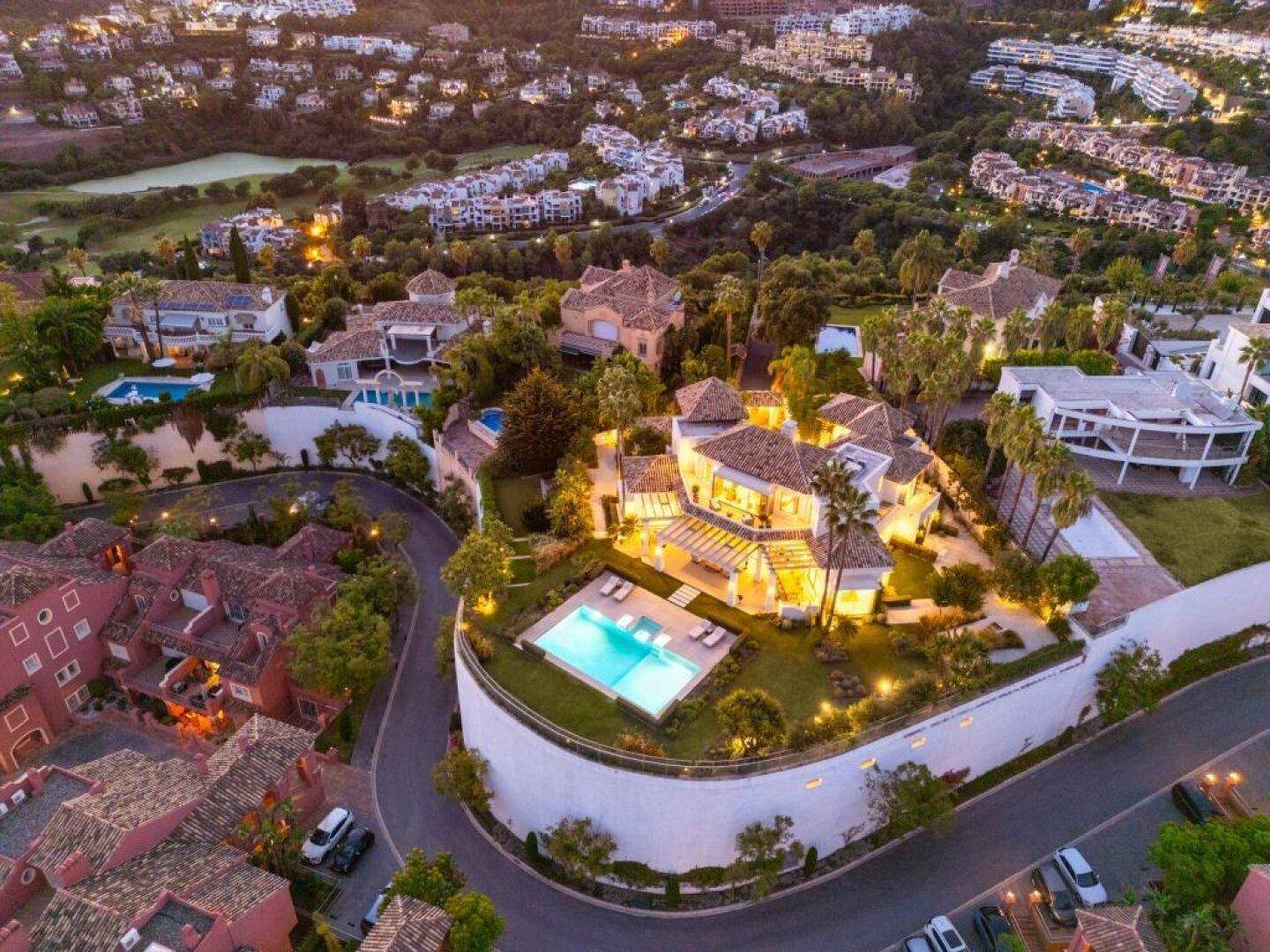 Weitläufige Luftaufnahme der beleuchteten Villa und der umliegenden Wohngegend bei Sonnenuntergang.