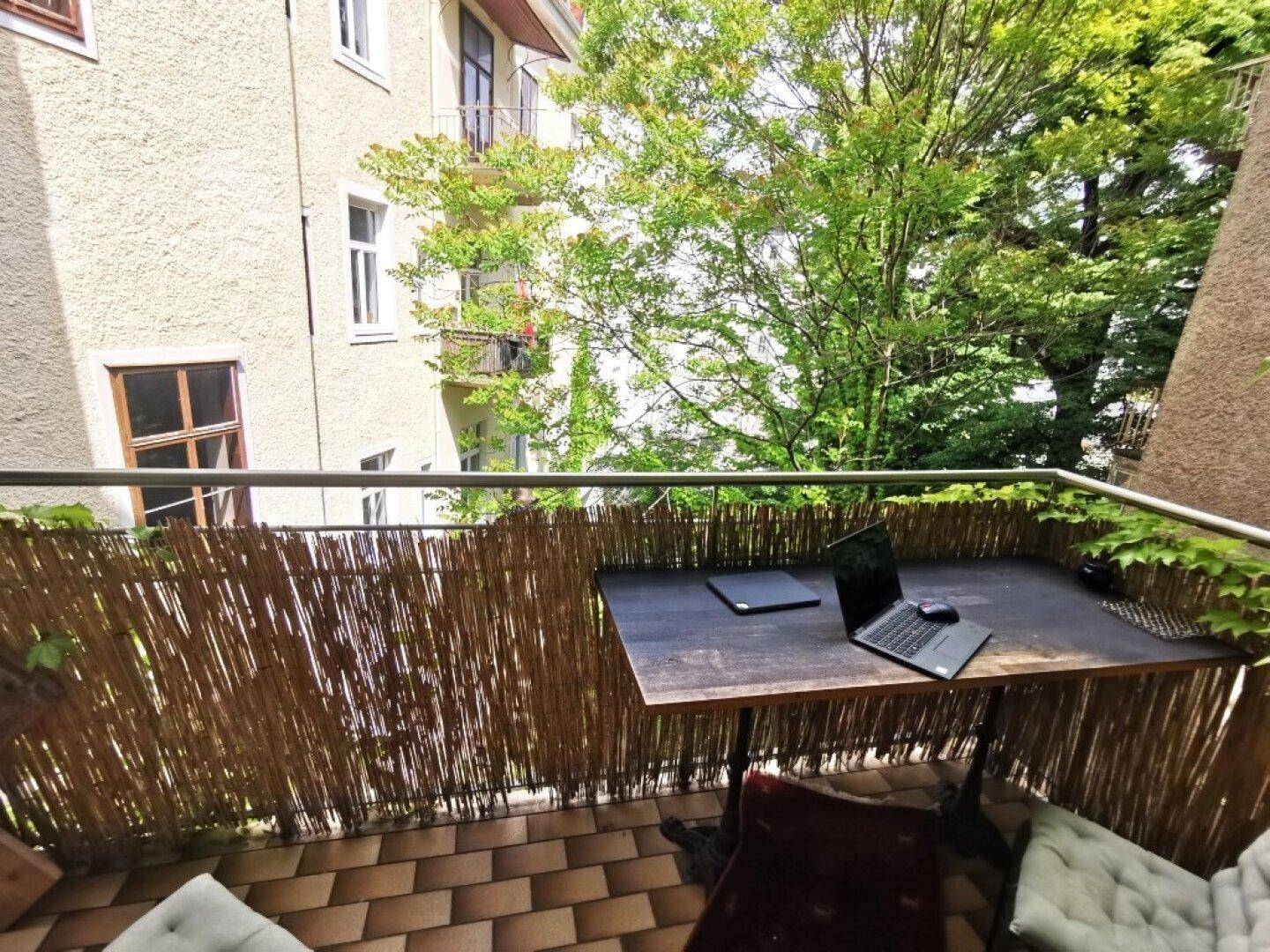 Gemütlicher Balkon mit Holztisch, Laptop und Blick auf grüne Bäume und das Nachbargebäude.