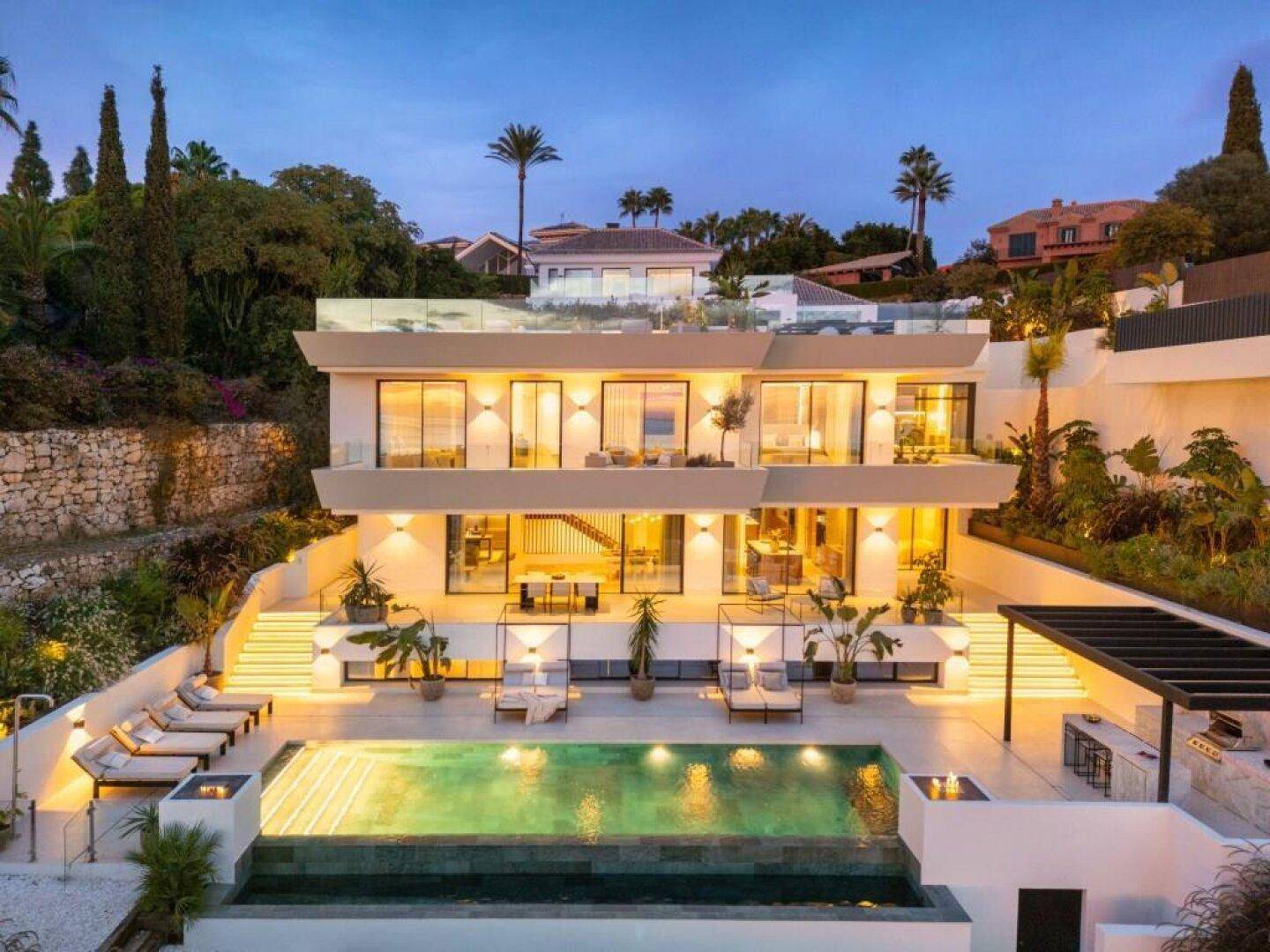 Elegante moderne Villa bei Dämmerung mit beleuchtetem Pool und mehreren Terrassen.