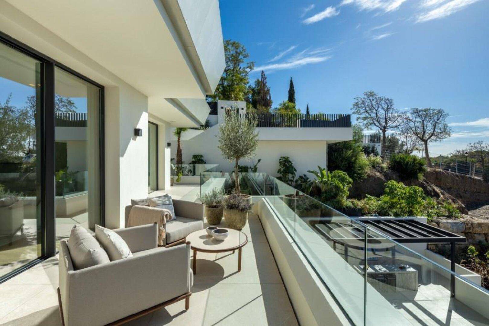 Sonniger Balkon mit Loungemöbeln, Glasgeländer und Blick auf die grüne Umgebung.