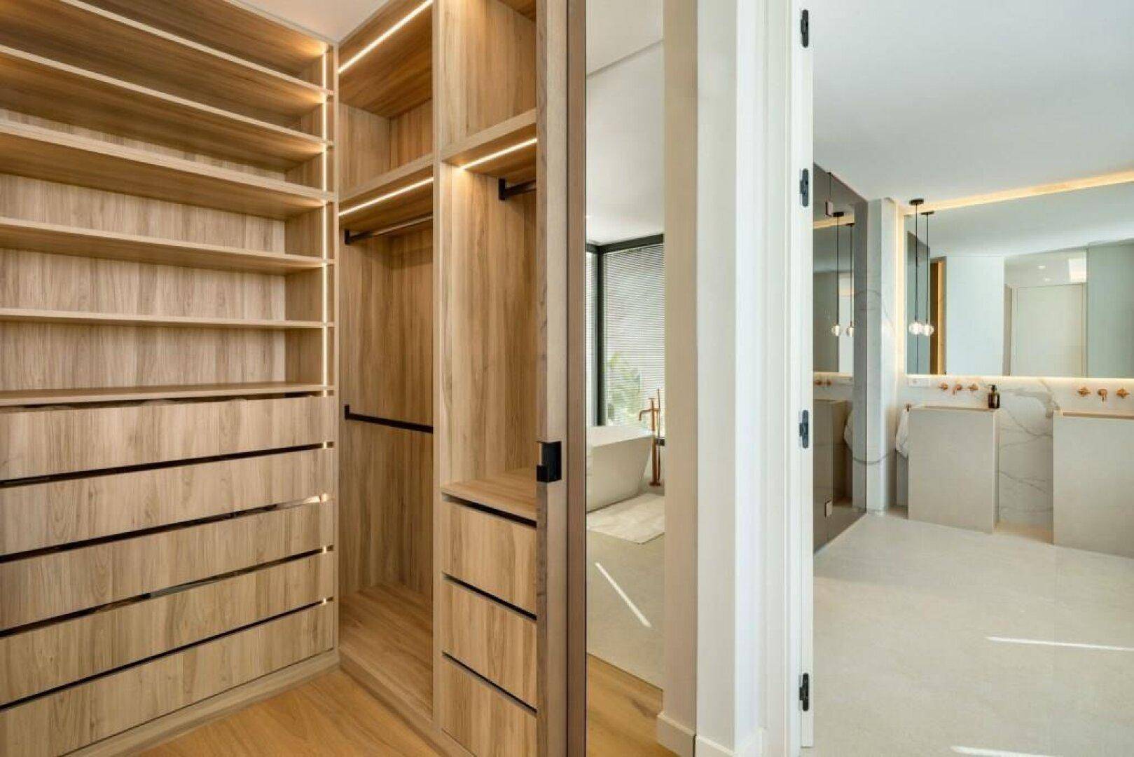 Geräumiger begehbarer Kleiderschrank mit Holzregalen und integrierter Beleuchtung, der zum Badezimmer führt.
