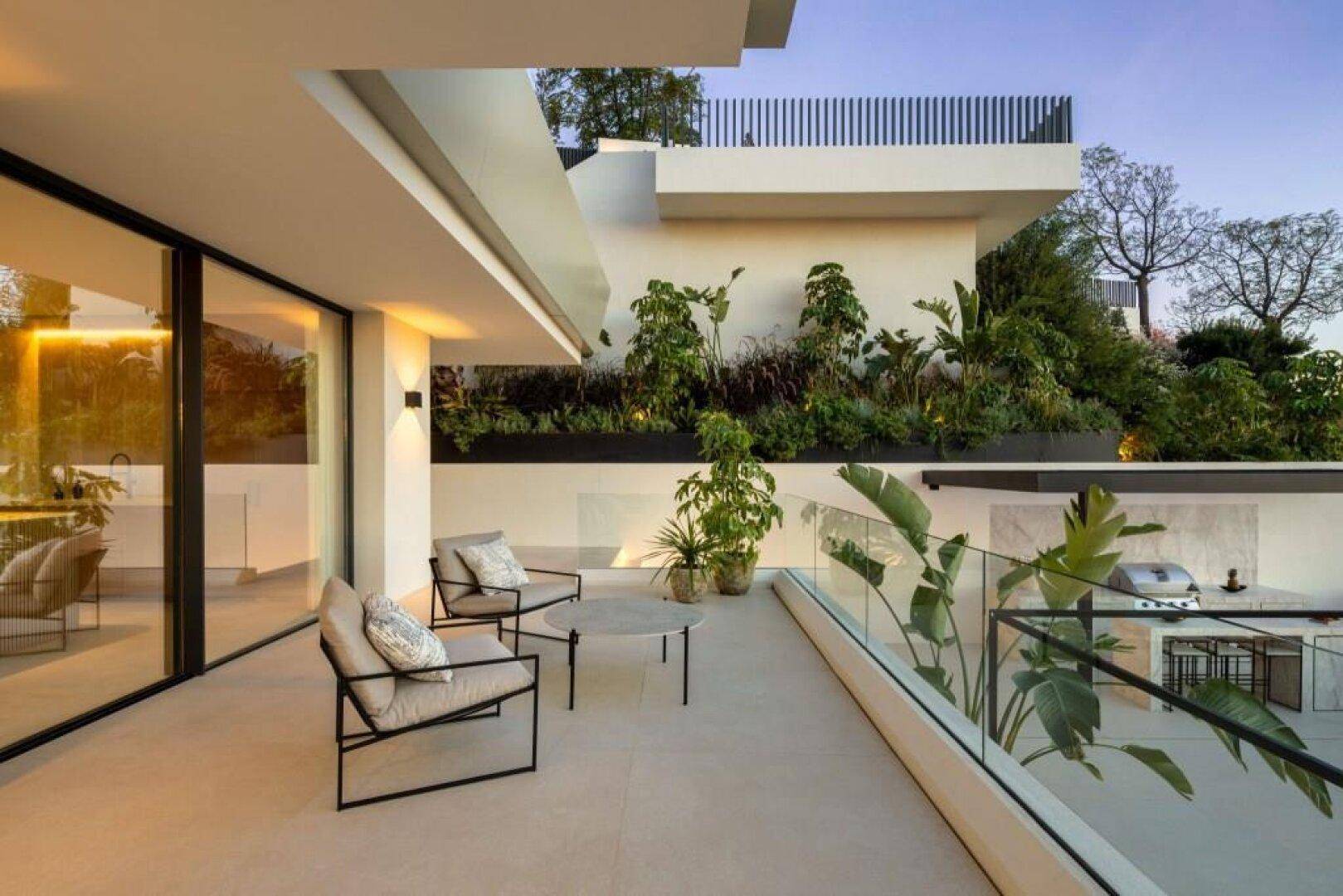 Moderne Terrasse mit minimalistischen Möbeln, großen Glasfronten und üppiger Bepflanzung für entspannte Stunden.
