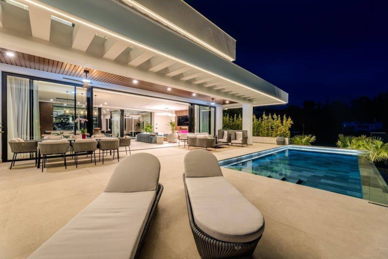 Stilvolle Terrasse bei Nacht mit Pool, Sonnenliegen und Essbereich im Freien.