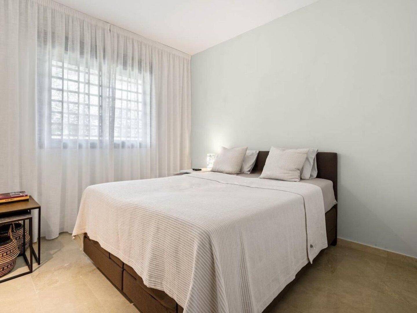 Helles Schlafzimmer mit Doppelbett, großen Fenstern und hellen Vorhängen, ausgestattet mit Fliesenboden.