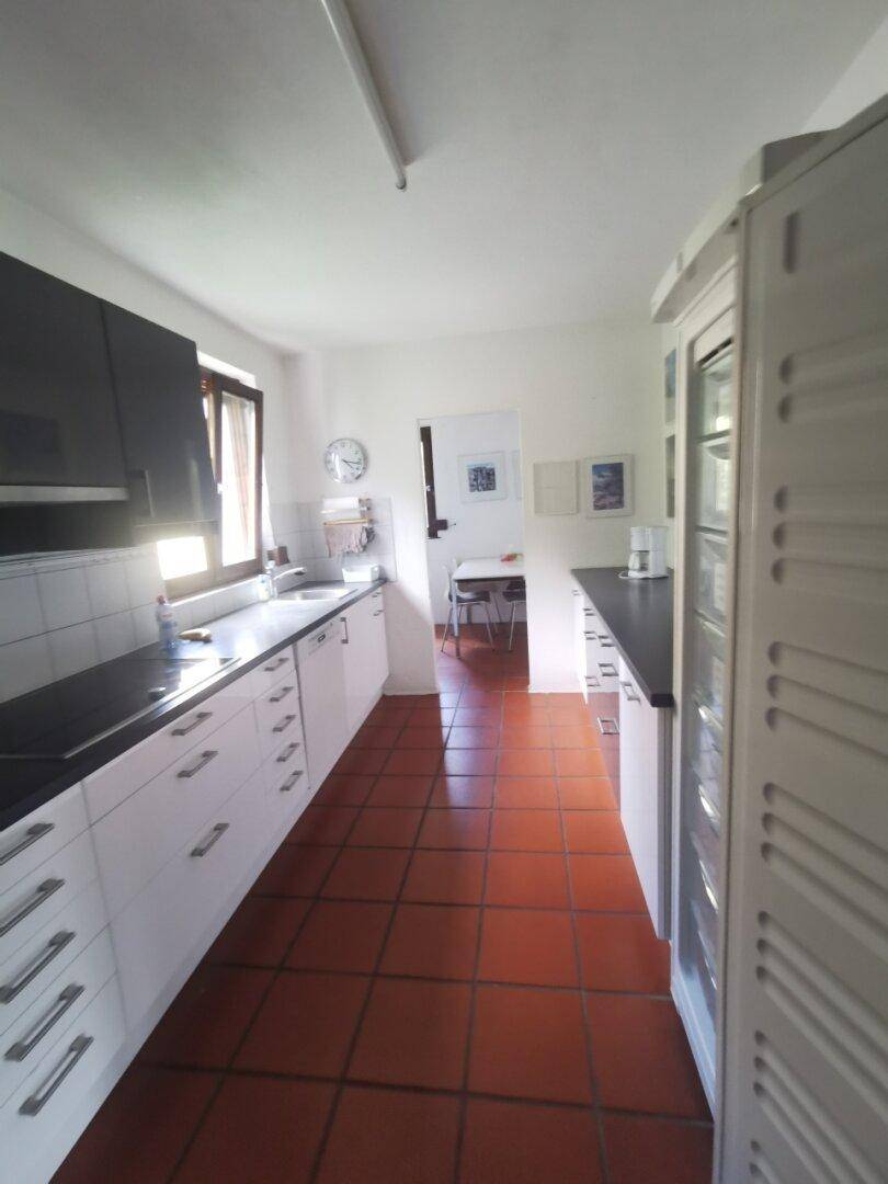 Lange Küche mit weißen Schränken, dunklen Arbeitsplatten und Terrakottafliesen, mit Fenster für Tageslicht.