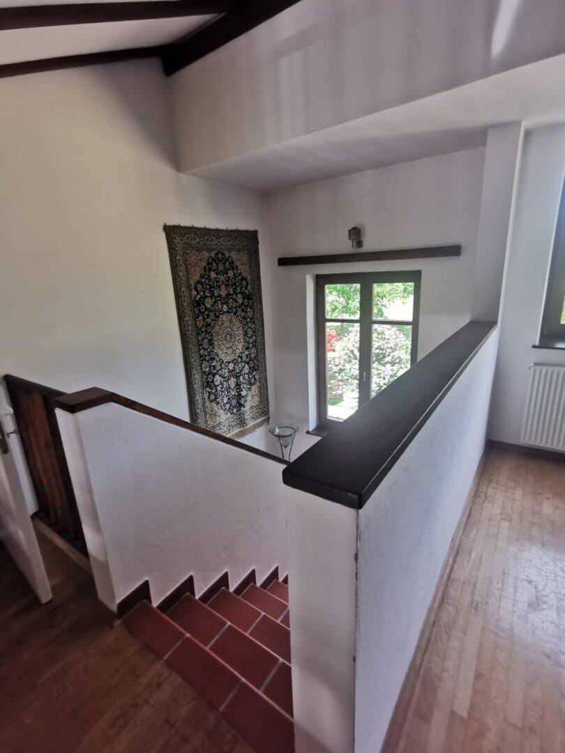 Treppenhaus mit Terrakottastufen, Holzgeländer und einem dekorativen Wandteppich, beleuchtet durch ein Fenster.