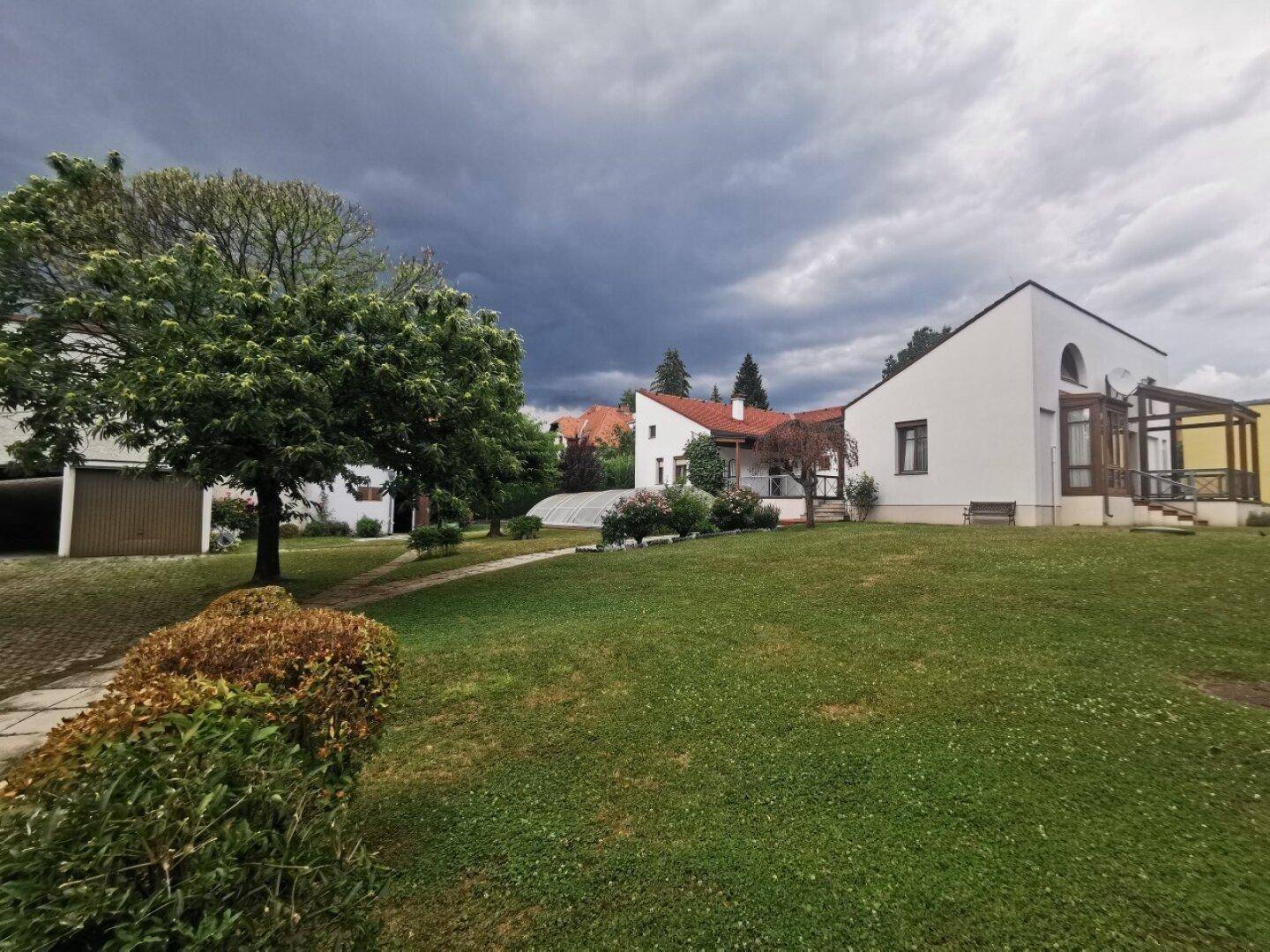 Weitläufiger Gartenbereich mit Rasenfläche, Bäumen und dem Wohnhaus mit überdachtem Pool unter Wolken.