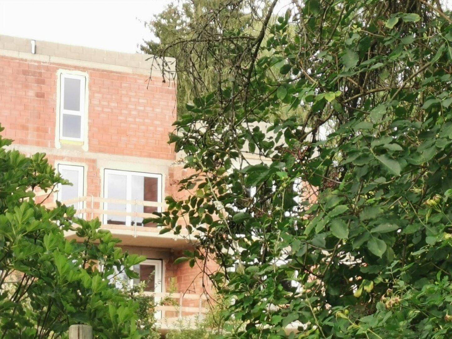 Teilansicht des Neubaus im Rohbau, umgeben von grünen Bäumen und Vegetation, mit sichtbaren Balkonen.