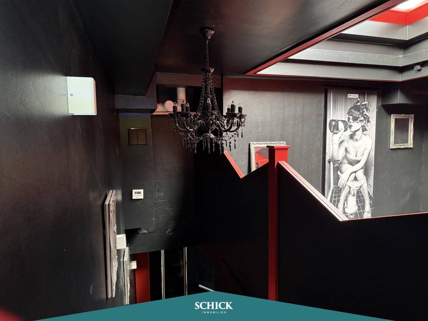 Blick von oben auf den Treppenbereich einer Bar mit dunklen Wänden und Kronleuchter.