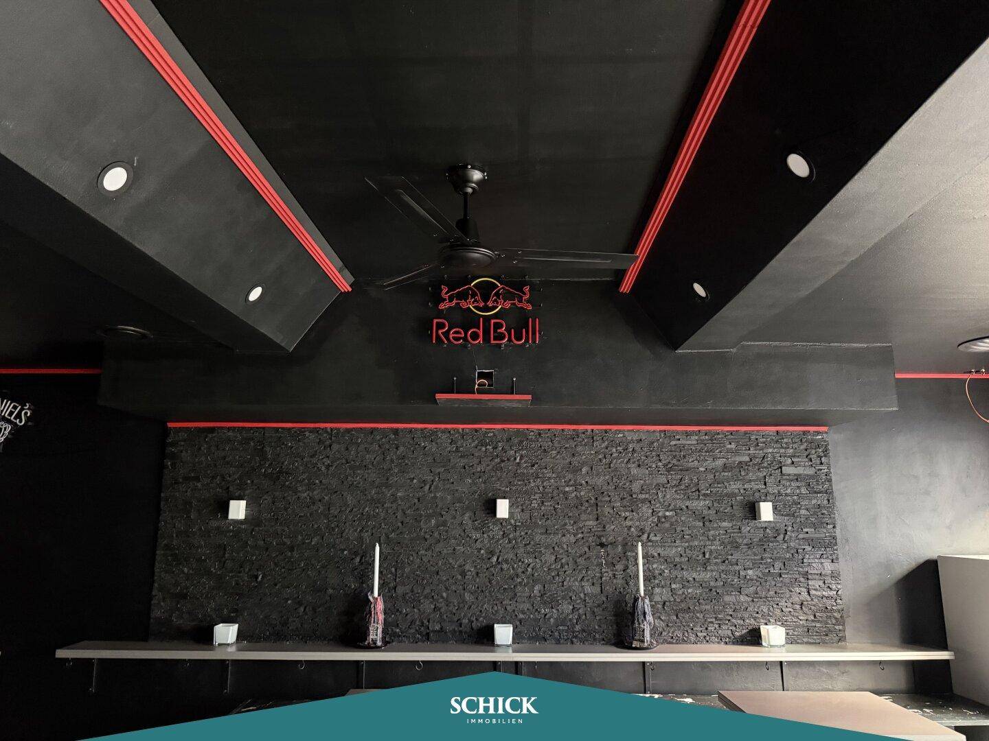Deckenansicht einer Bar mit schwarzem Ventilator, roten Lichtleisten und dunkler Steinwand.