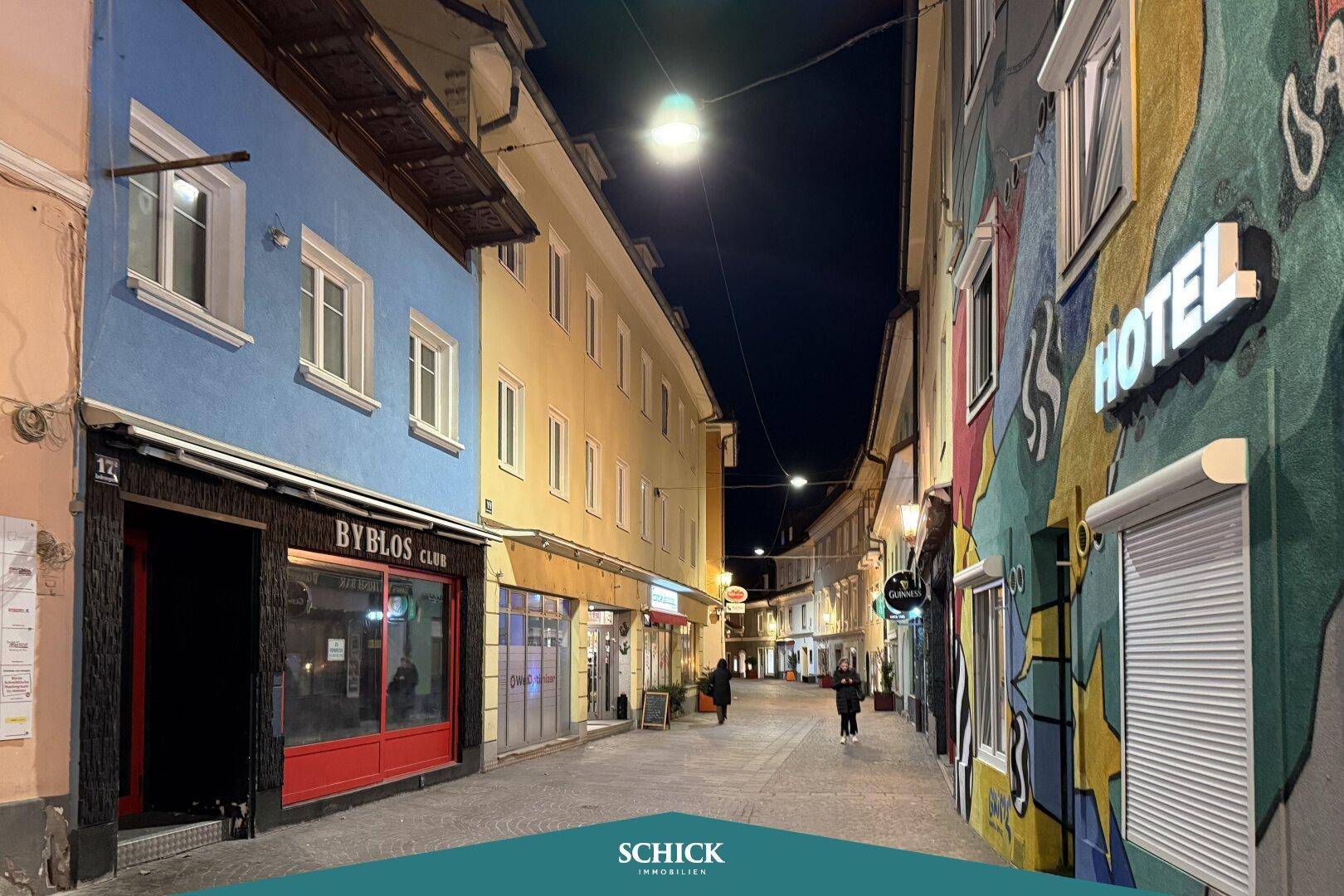 Nachtansicht einer belebten Gasse mit verschiedenen Gebäuden und Geschäften.