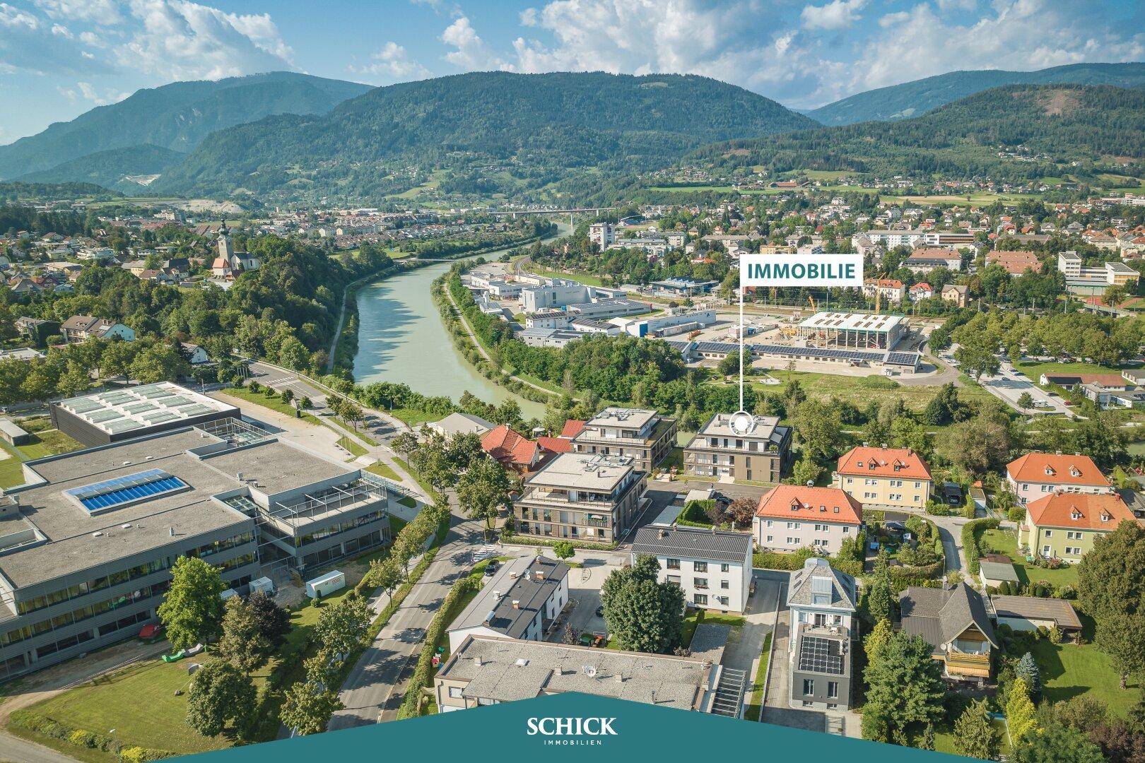 Weitläufige Ansicht des Gebäudekomplexes in städtischer Umgebung mit Fluss und Bergpanorama.