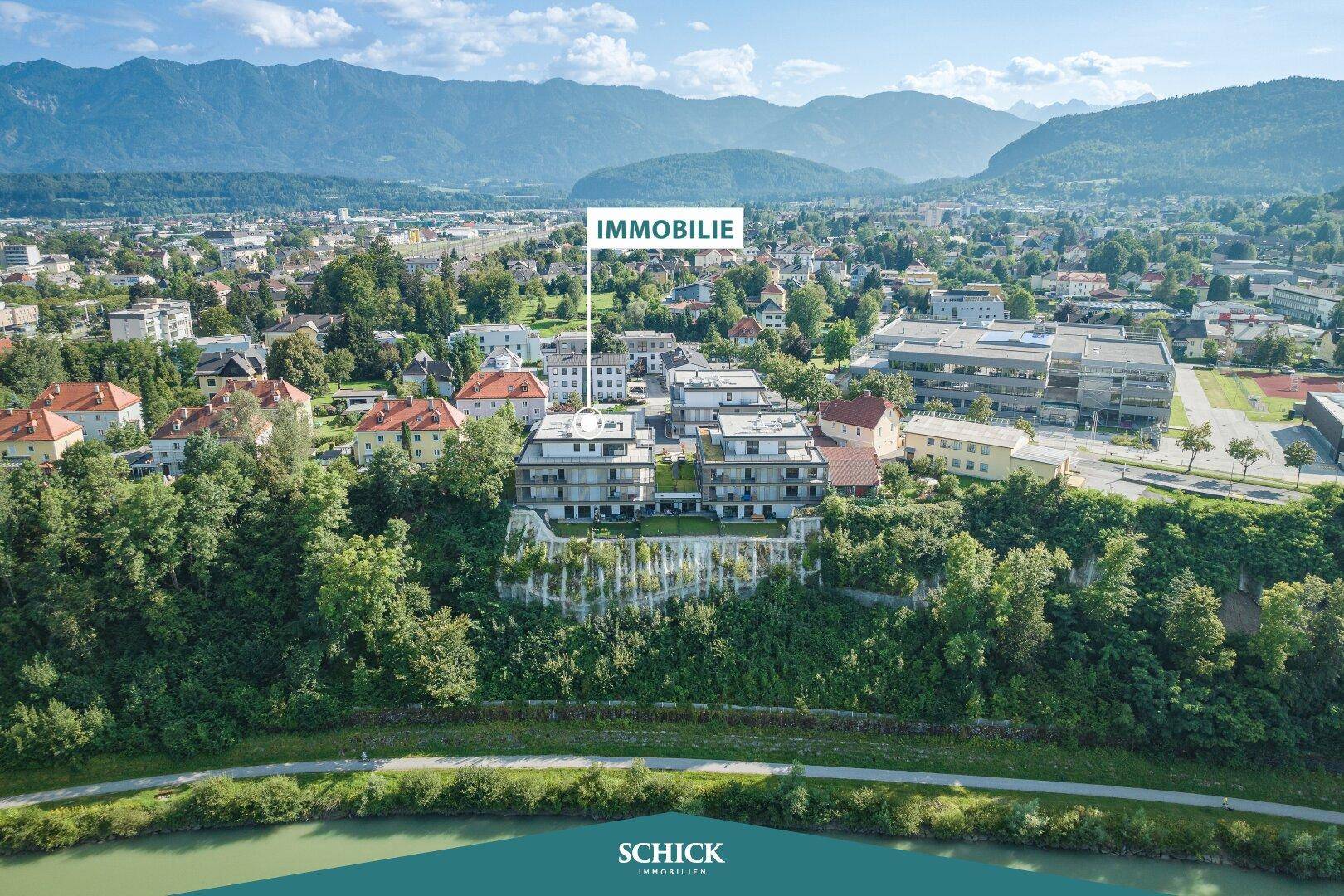 Panoramablick auf den Gebäudekomplex am Flussufer mit Bergen im Hintergrund und grüner Umgebung.