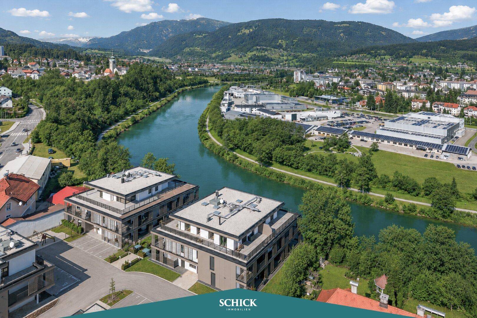 Luftaufnahme von modernen Wohngebäuden am Flussufer mit umliegender Stadt- und Berglandschaft.