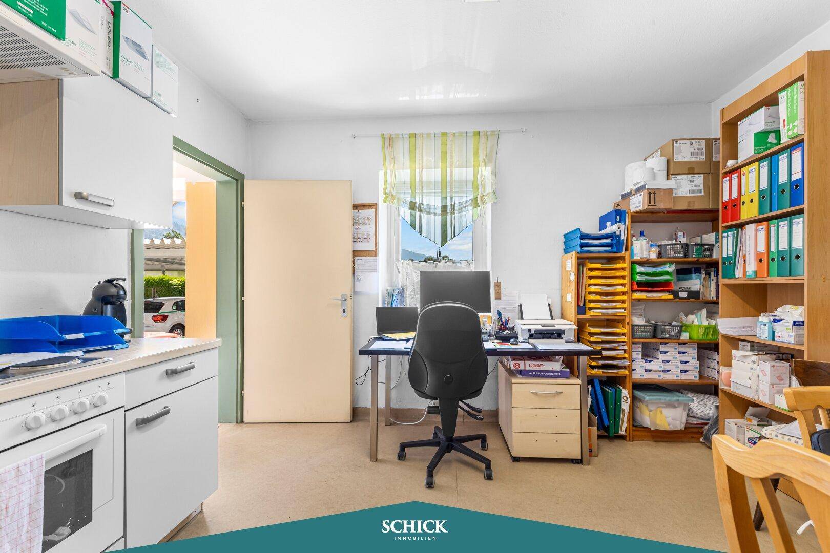 Geräumiges Büro mit Schreibtisch, Regalen und einer kleinen Küchenzeile im Hintergrund.