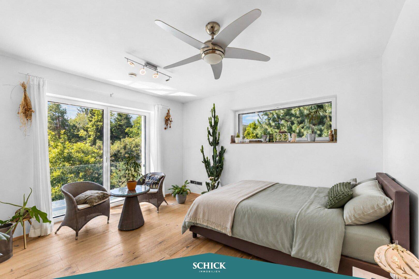 Helles Schlafzimmer mit Deckenventilator, Sitzgelegenheit und großen Fenstern mit Blick ins Grüne.