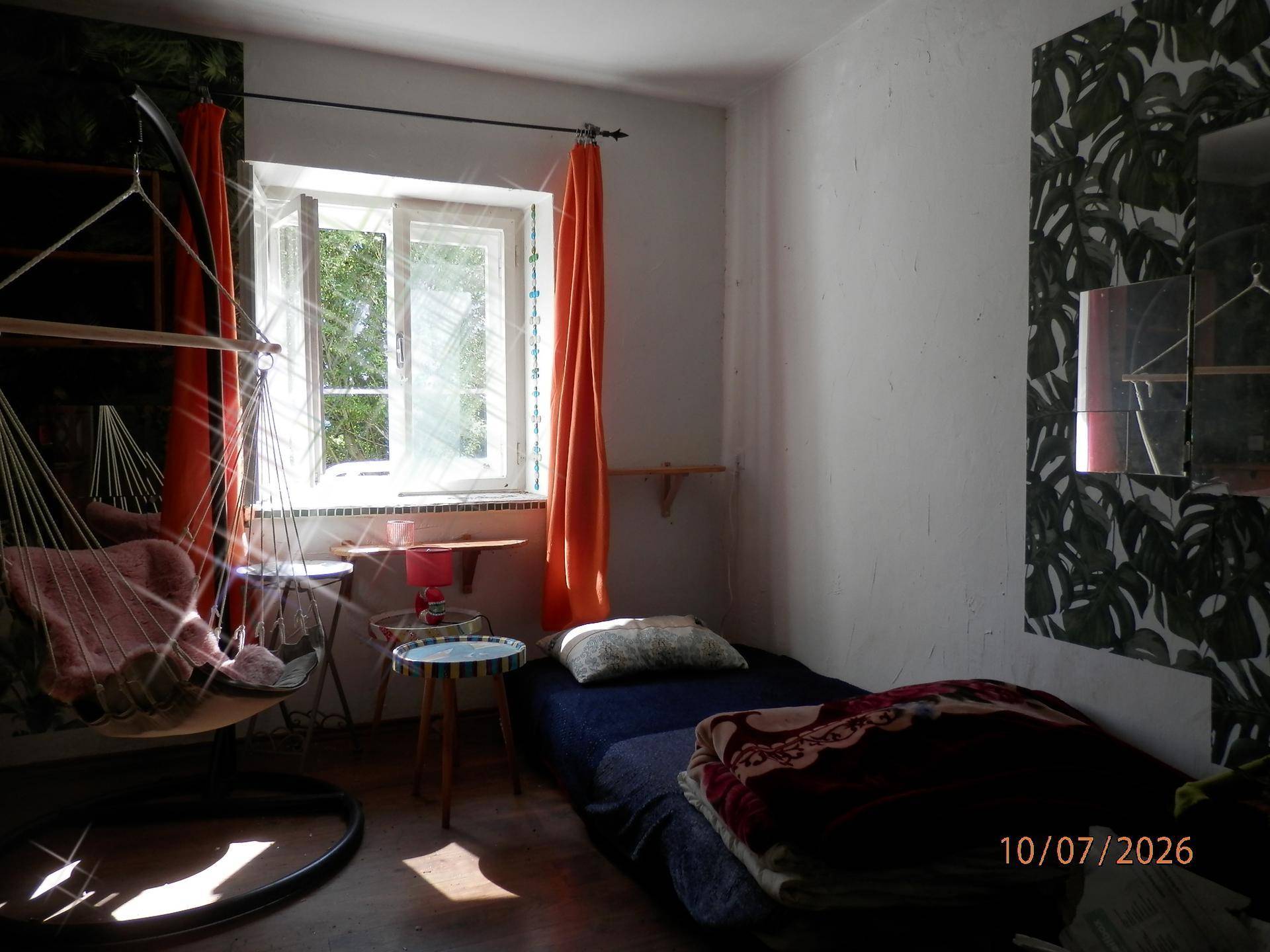 Helles Schlafzimmer mit Einzelbett, Hängesessel und orangefarbenen Vorhängen.