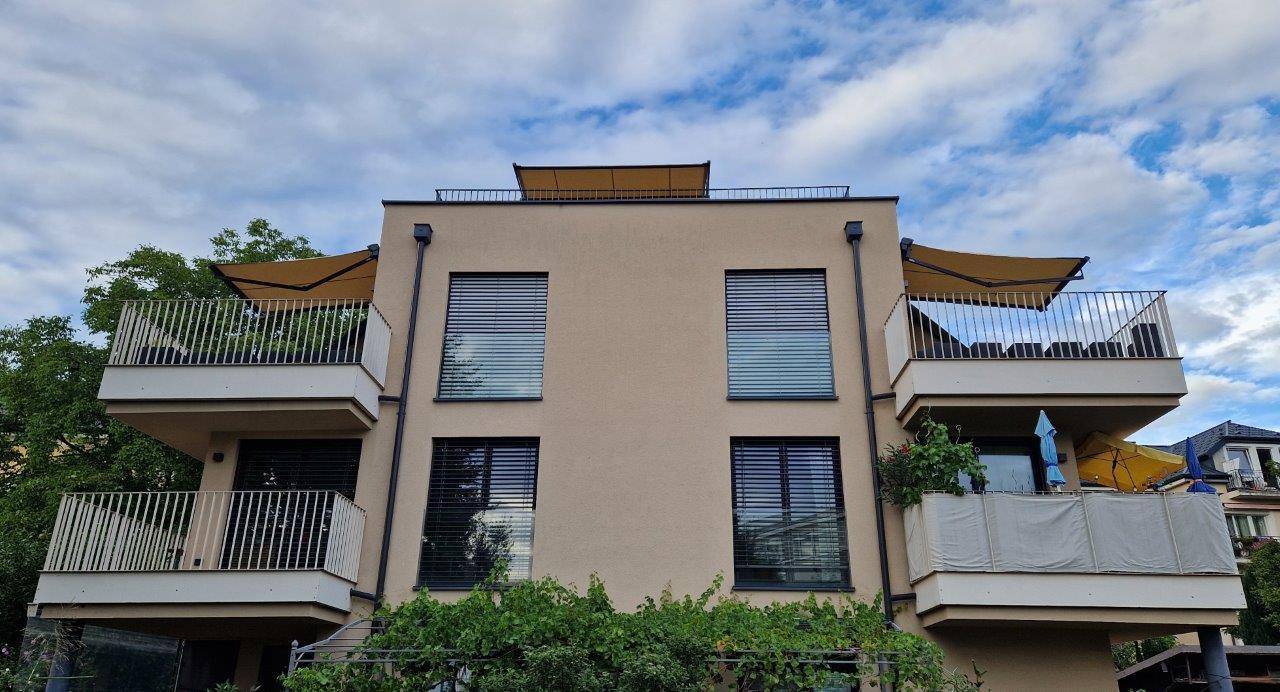 Modernes Wohngebäude mit ausgefahrenen Markisen über den Balkonen und beiger Fassade.
