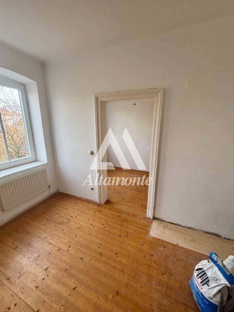 Einfaches Zimmer mit Holzboden und Türöffnung zu einem weiteren Raum, hell und funktional.