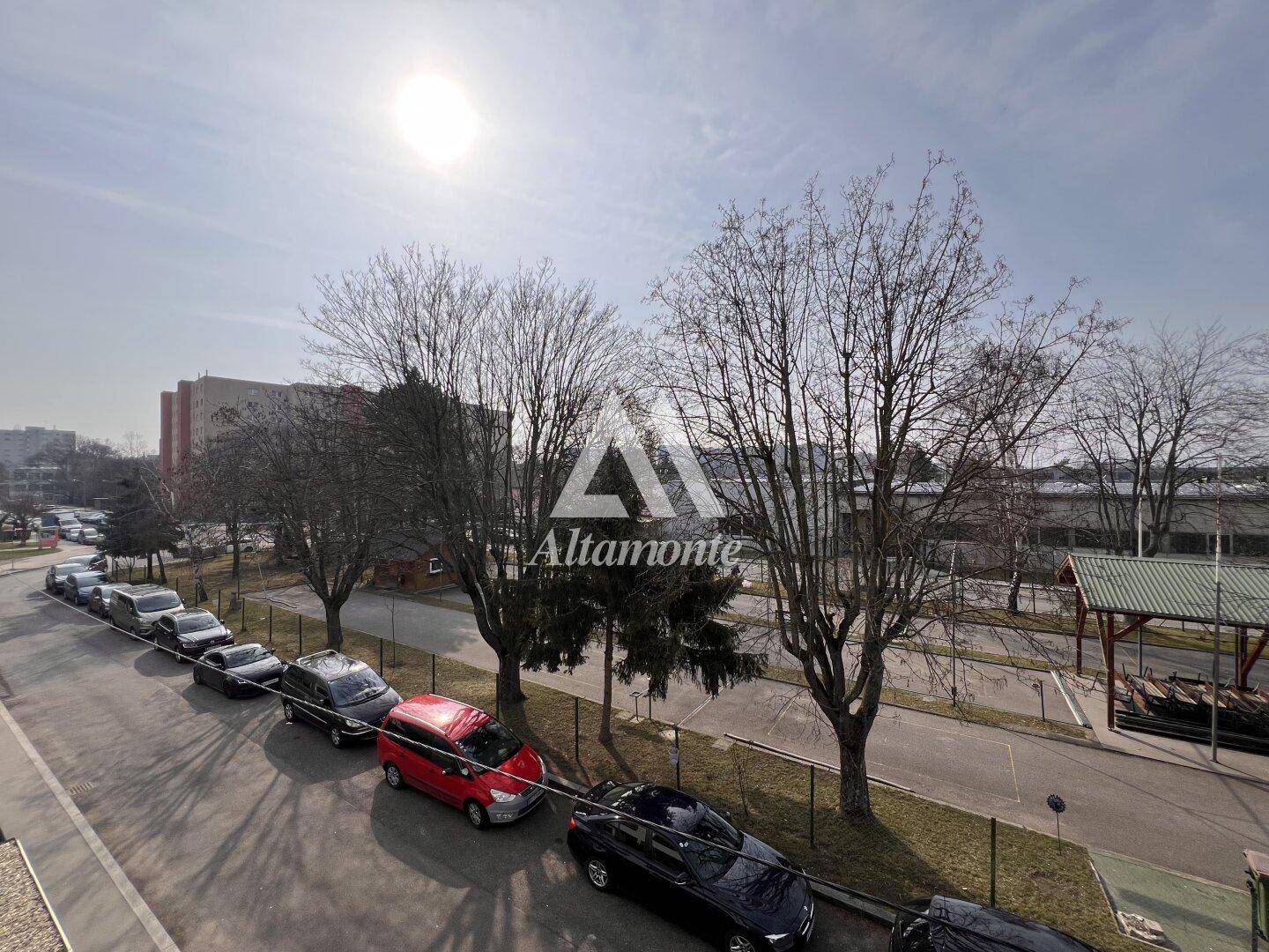 Sonnenbeschienener Blick auf die Straße mit parkenden Autos und Bäumen, umgeben von städtischer Bebauung.
