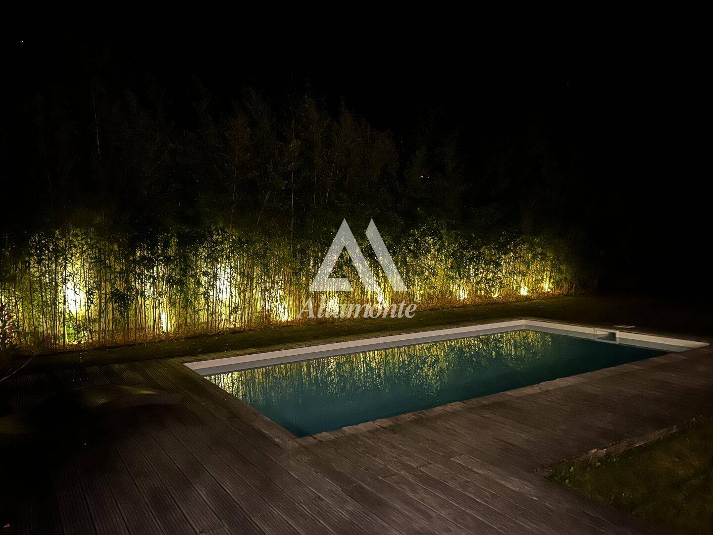 Beleuchteter Poolbereich bei Nacht, umgeben von Bambuspflanzen und Holzdeck.